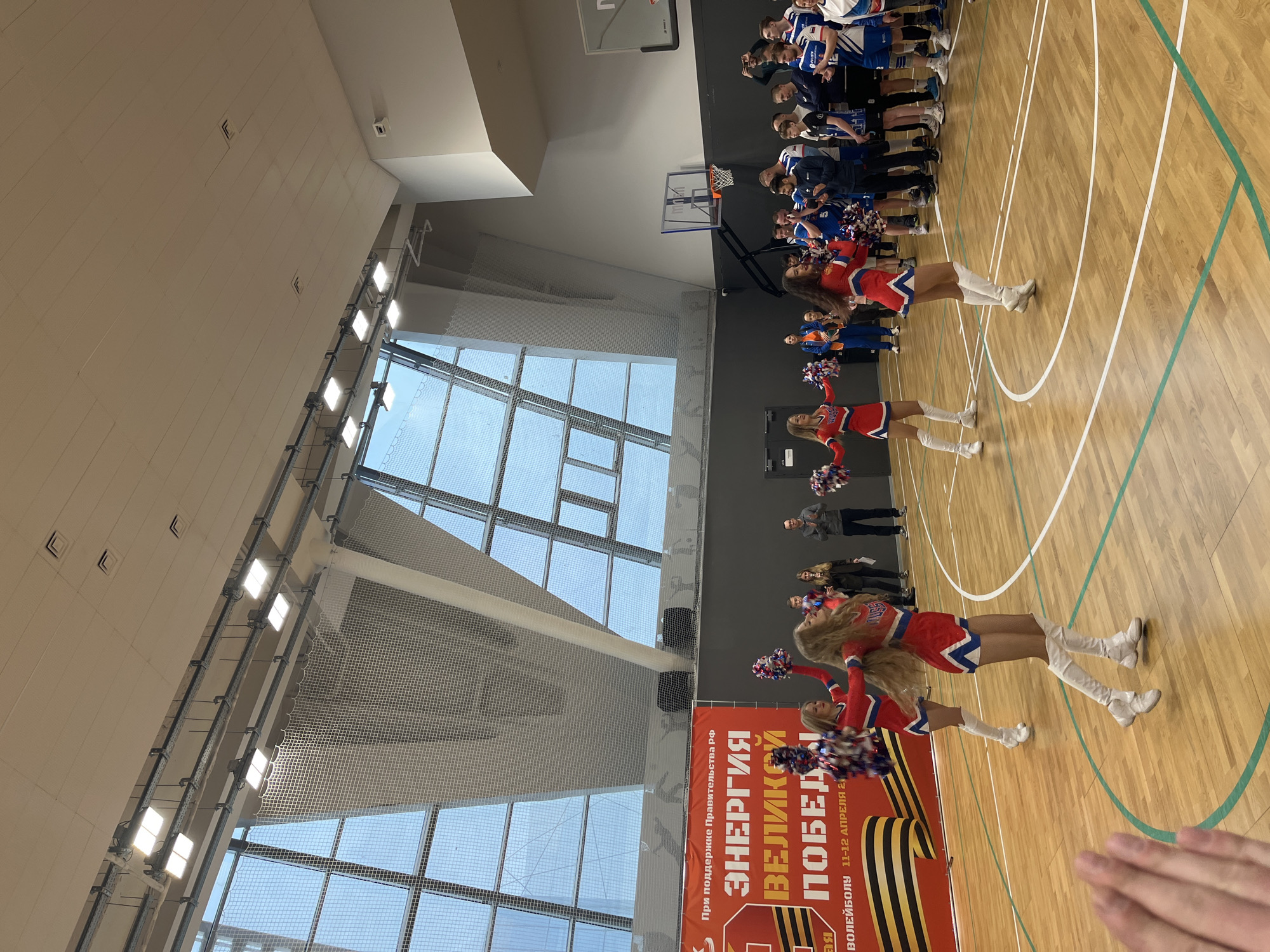 The image depicts a basketball court inside a sports hall in Moscow, Russia. Cheerleaders in red and white uniforms with white boots stand along the court, holding pom-poms. A basketball team in blue and white uniforms stands in a line near the basketball hoop. A large sign with Russian text, "ЭНЕРГИЯ ВЕЛИКОЙ ПОБЕДЫ" (Energy of Great Victory), is visible. Several spectators are standing in the background. The hall has a high ceiling with multiple rows of lights. A large window is visible, with a net covering it. The floor is wooden with painted lines marking the basketball court.