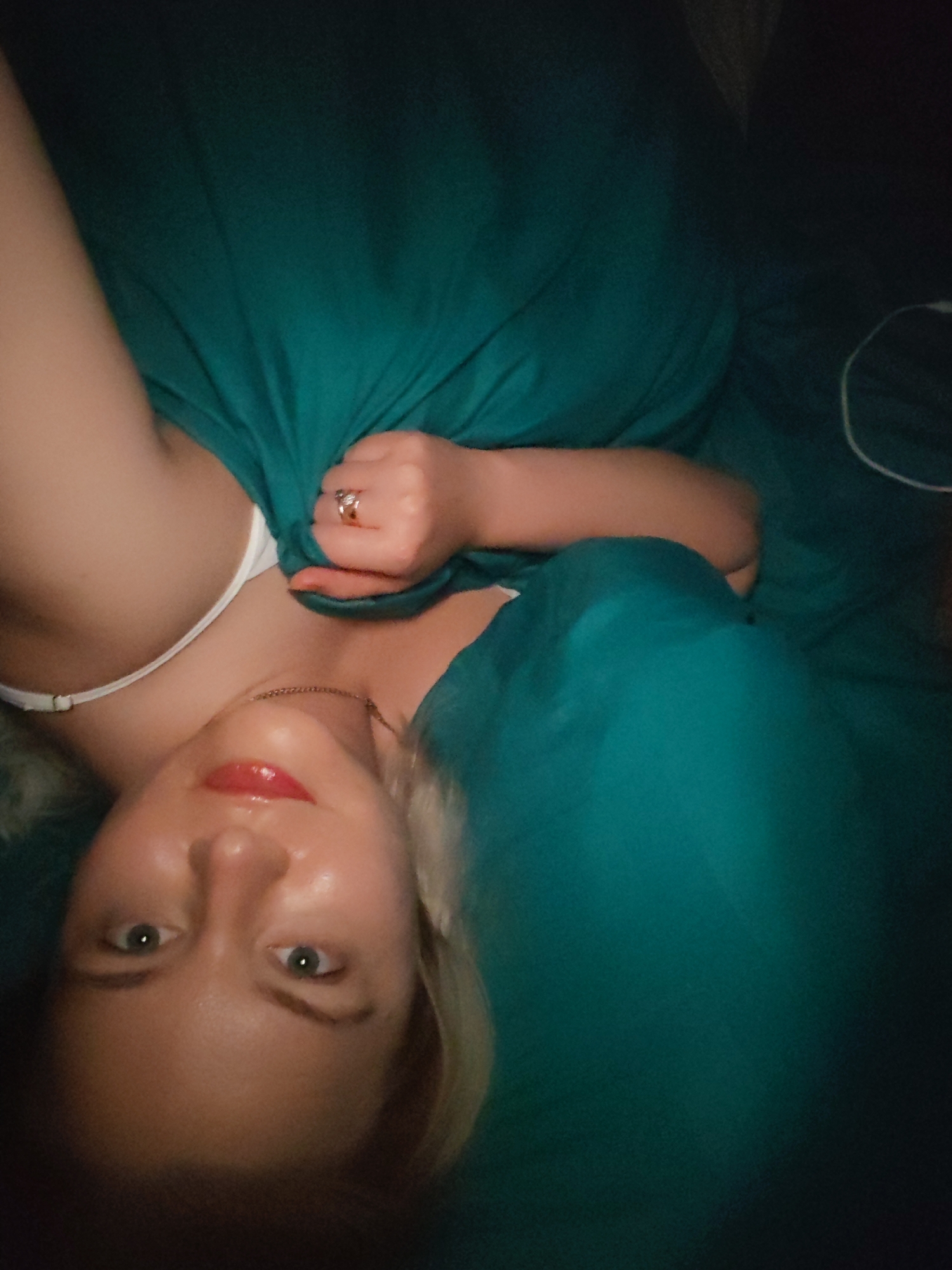 A woman lies on a teal-colored surface, possibly a bed, with her head towards the bottom of the frame. She is fair-skinned with blonde hair and green eyes. She is wearing a white top with thin straps and a gold necklace. She has red lipstick on. Her right hand is visible, clenched around a piece of teal fabric, and she is wearing a gold ring. A white cord is visible in the upper right corner of the frame. There is no evidence to suggest the location is in Izhevsk, Russia.