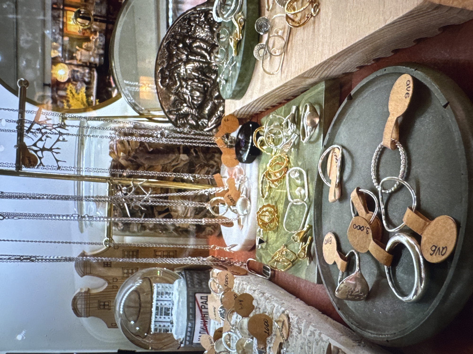 The image shows a display of jewelry, predominantly rings and necklaces, in what appears to be a shop or market stall. The items are arranged on various surfaces, including a rough, textured white stand, a green tiered display, and a dark circular tray. Several pieces of jewelry are tagged with small, brown circular labels, some of which have handwritten numbers or letters. In the background, a snow globe with a miniature building inside is visible, alongside wooden decorative houses and a jar filled with dried organic material. A round mirror reflects another area of the shop, showing more display items and lighting. The overall impression is of a curated collection of artisanal jewelry. There are no people visible in the image. The location "Новинки, Russia" is not discernible from the visual information presented in the image.