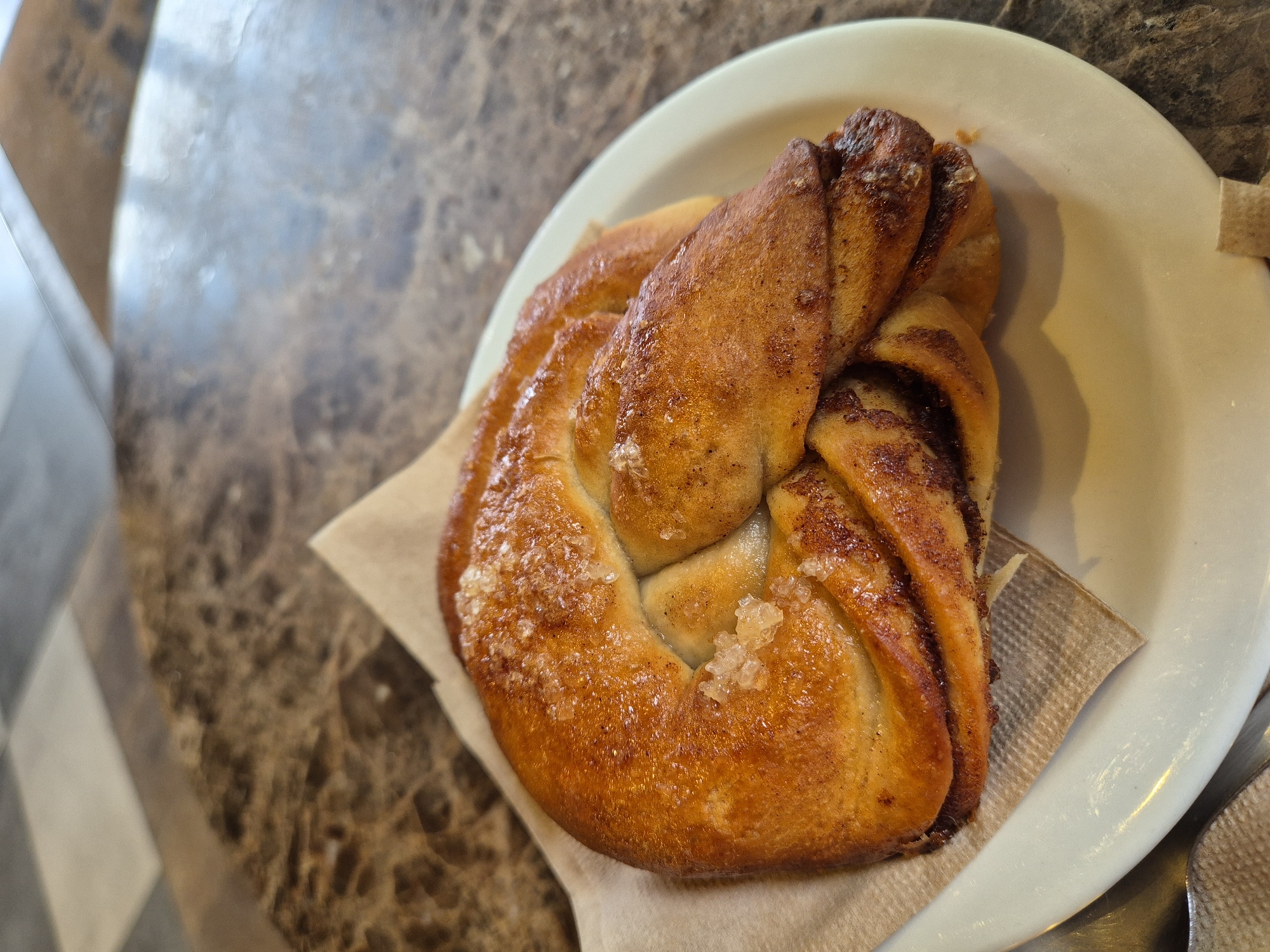 A close-up shot of a cinnamon bun on a small, off-white plate lined with a light brown paper napkin. The bun, glazed and sprinkled with large sugar crystals, is served on a marble-patterned table. A portion of the checkered floor is visible in the background. The bun rests on a table in West Suffolk, United Kingdom.