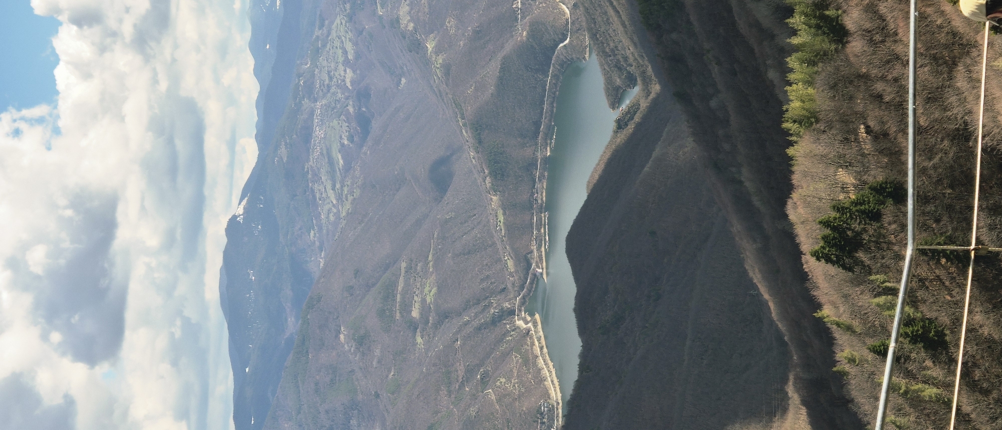 An aerial view, possibly from a gondola, cable car, or similar structure (metal poles and cables are visible), shows a landscape with mountains, a long, narrow lake or reservoir, and forests in Ravnogor, Bulgaria. The mountains are predominantly covered with dry, bare-branched trees and some areas of sparse green vegetation. Snow traces are visible on higher mountain peaks. The lake stretches through a valley, with roads running alongside at least one edge. The sky is partly cloudy with blue patches.