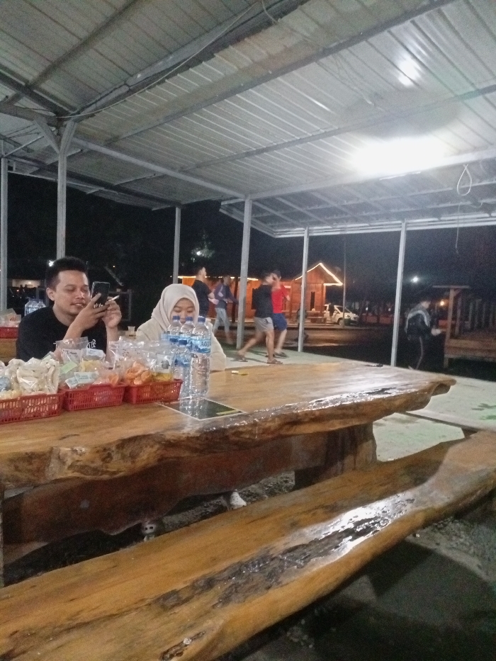 This image depicts a nighttime scene in an open-air or semi-outdoor dining area, likely in Pangalengan, Indonesia. A sturdy metal roof structure covers the area, which is illuminated by artificial lights, casting reflections on the roof.
In the foreground, two people are seated at a large, rustic wooden table. The table is a thick slab of wood with natural, uneven edges, accompanied by a matching wooden bench. On the left, a man with short, dark hair, wearing a black t-shirt, is holding a smartphone in his left hand and what appears to be a cigarette or vape in his right. He is looking intently at his phone. To his right, a woman wearing a light-colored hijab is also seated, looking down towards the table.
On the table in front of them are several red plastic baskets filled with various packaged snacks, such as crackers and chips. Multiple plastic bottles of "AQUA" branded mineral water are also visible.
In the mid-ground, several blurry figures of people can be seen standing and walking, suggesting a casual gathering or an active public space. One person in a red shirt is noticeable among them. Further in the background, some structures with illuminated outlines, possibly small wooden cabins or buildings, are visible against the dark night sky, hinting at a recreational area or a resort. The overall atmosphere appears relaxed and social.