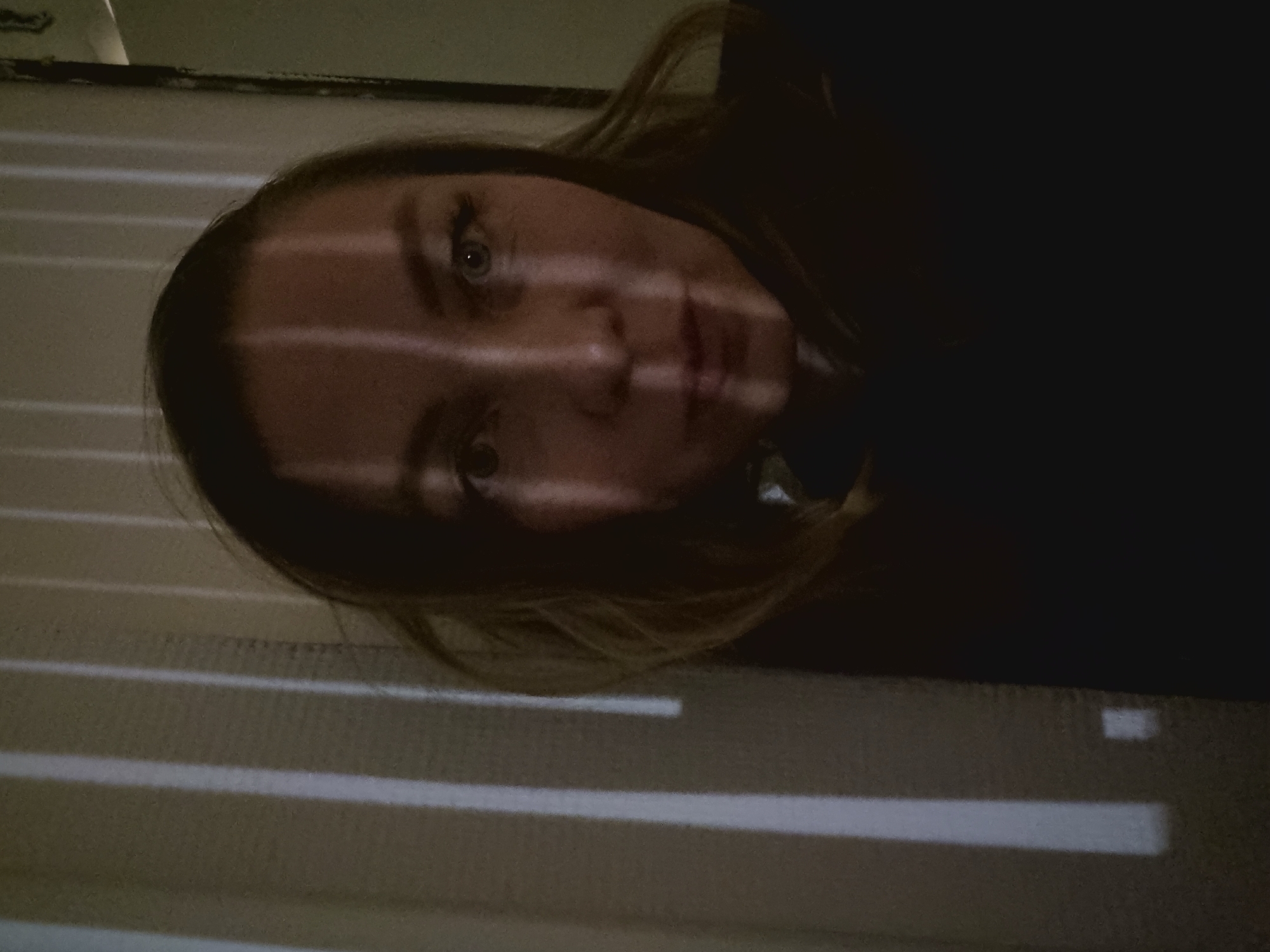 A low-light photograph depicts a person with light skin and light brown hair looking directly at the camera. Horizontal bands of bright light illuminate the face, creating a striped effect. The person's eyes are visible and appear to be light-colored. The right side of the face is partially obscured by shadow. The background consists of more horizontal light bands above and below the person's face. The setting and specific location in Kemul, Russia are not identifiable from the image.