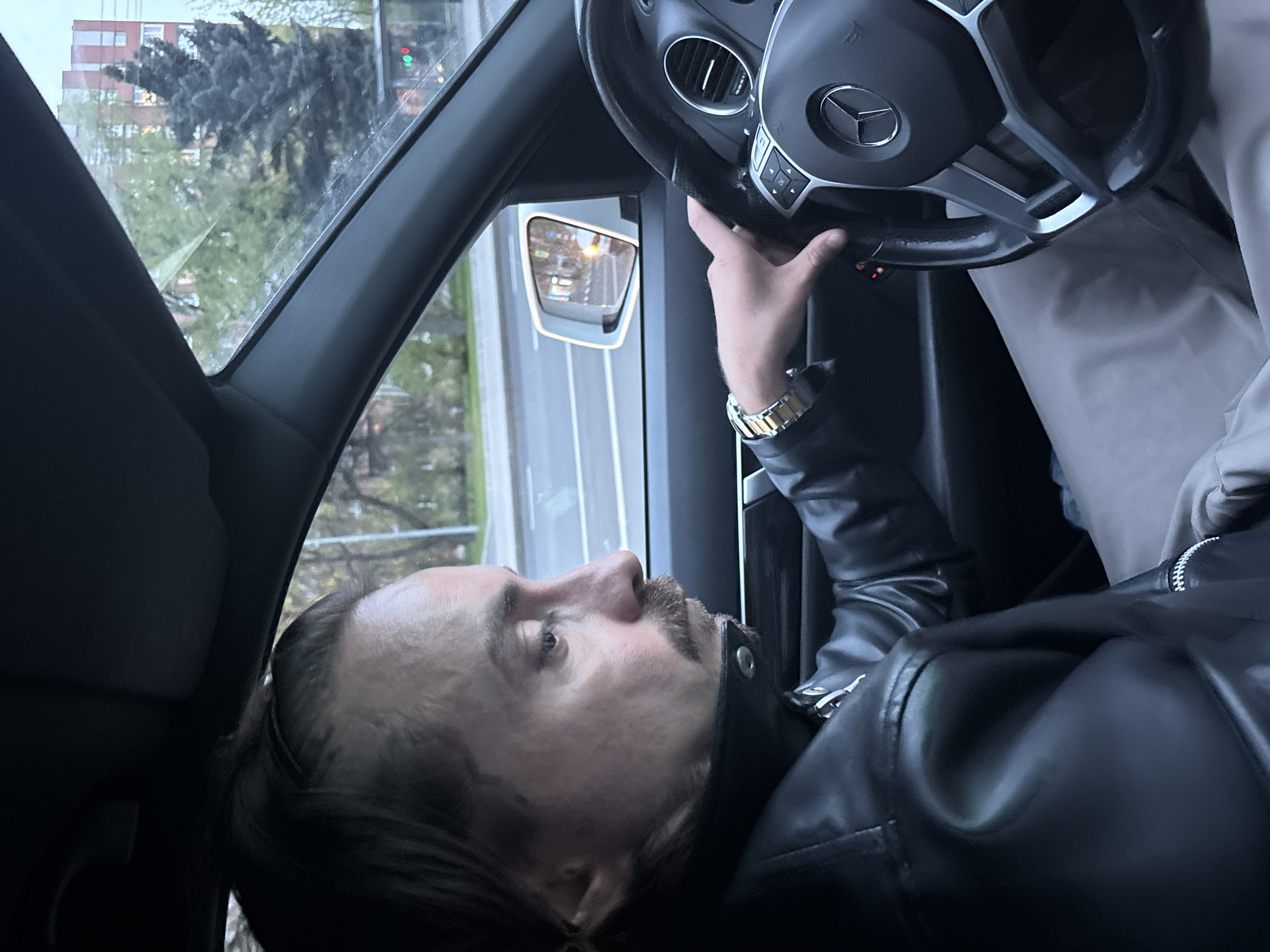 The image shows a man in the driver's seat of a car, holding the steering wheel. The car is a Mercedes-Benz. The man is wearing a black leather jacket and a gold watch. Outside the car window is a view of a street in Saint Petersburg, Russia. There is a building and a traffic light visible. The traffic light is green.