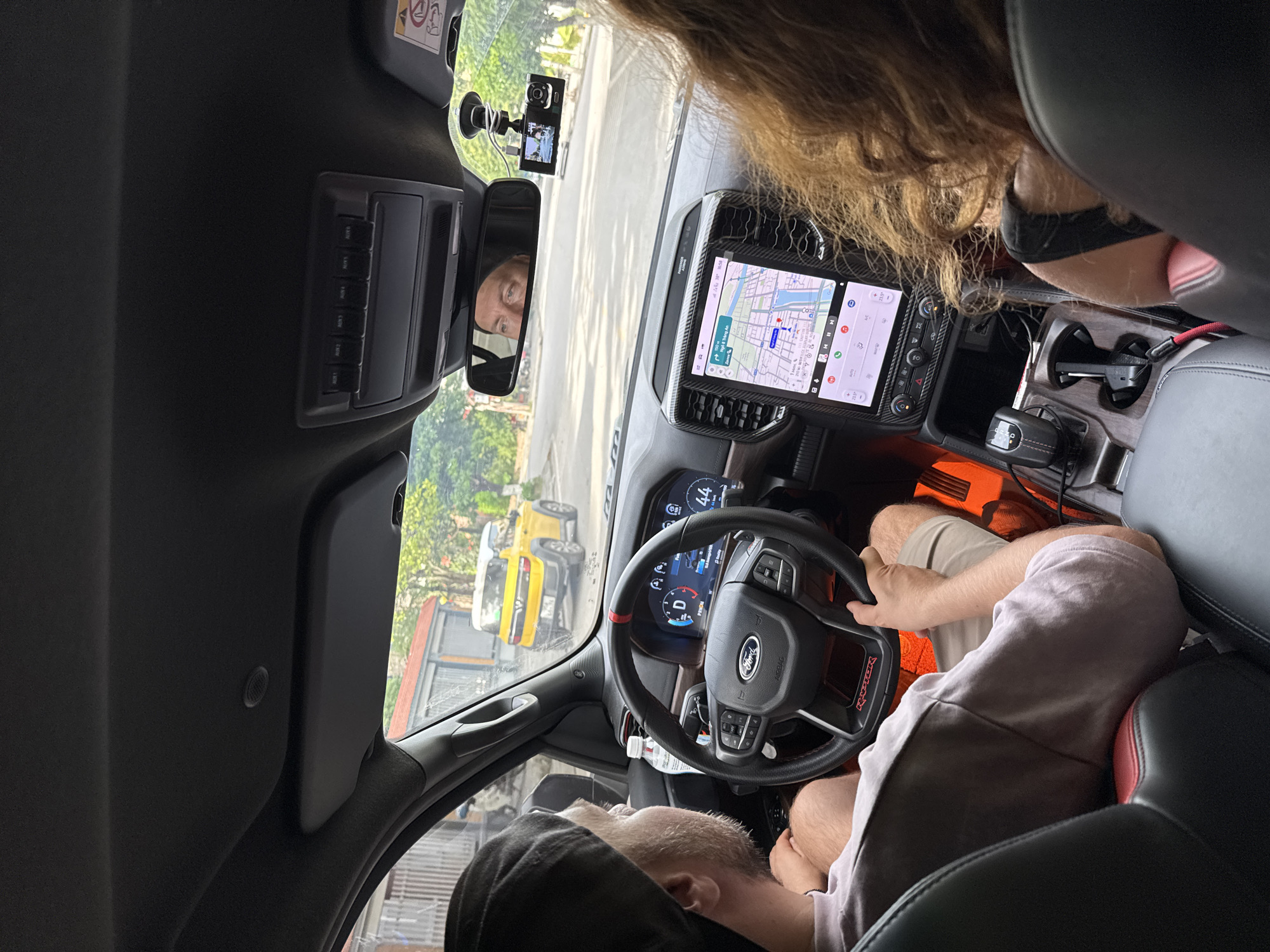 The image shows the interior of a Ford Ranger Raptor pickup truck, likely in Hoa Lư Ward, Vietnam, based on the surrounding environment. A man wearing a black baseball cap and a light-colored t-shirt is driving. In the passenger seat, a woman with long, wavy brown hair is visible, leaning forward. The truck's dashboard features a large touchscreen display showing a navigation map, a digital instrument cluster, and various controls. A dashcam is mounted on the windshield. Outside the vehicle, a yellow vehicle is visible on a street lined with buildings and trees. The overall scene depicts a moment of travel within a Vietnamese urban or suburban setting.