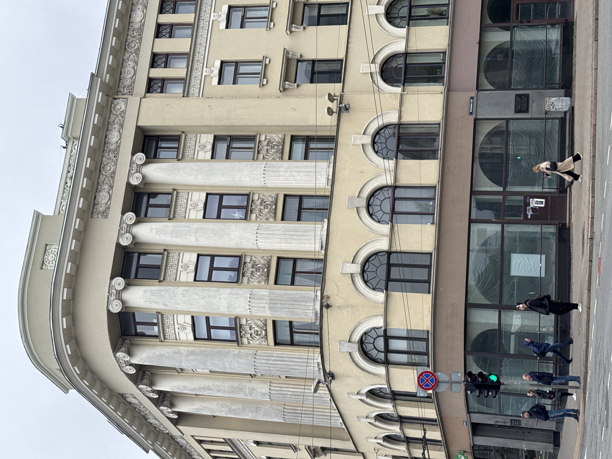 The image shows the facade of a building in Riga, Latvia, featuring architectural details such as columns with Corinthian capitals, rectangular and arched windows, and decorative elements. On the street level, there are large windows, a doorway, a traffic light showing green, and a red and blue no-stopping sign. Several pedestrians are visible on the sidewalk, including one woman walking towards the building's entrance and a group of people crossing the street.