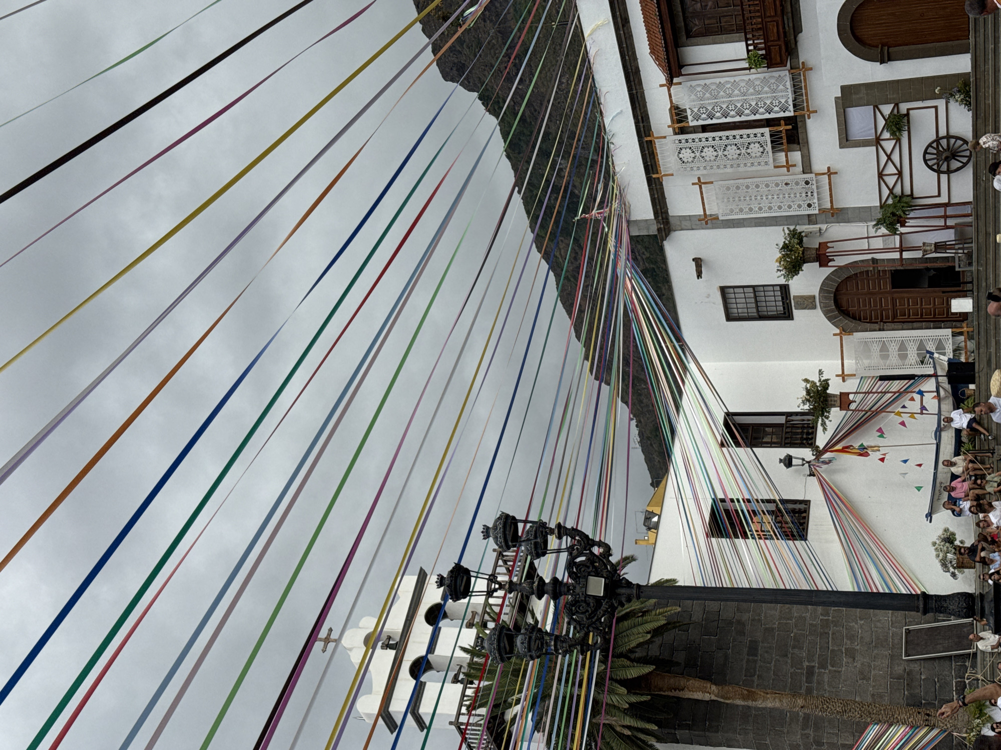 In an outdoor plaza, a multitude of colorful ribbons are strung across the sky, creating a festive canopy. Below, a white-walled building with traditional architecture features arched doorways, windows with wooden shutters, and decorative white lace hangings. A church bell tower with a cross stands to the left, next to a tall palm tree. A black, ornate lamppost is positioned in the foreground. Several people are seated on steps in front of the building, and a small white boat rests on a platform to the right. In the background, a dark, lush mountain rises against a cloudy sky.