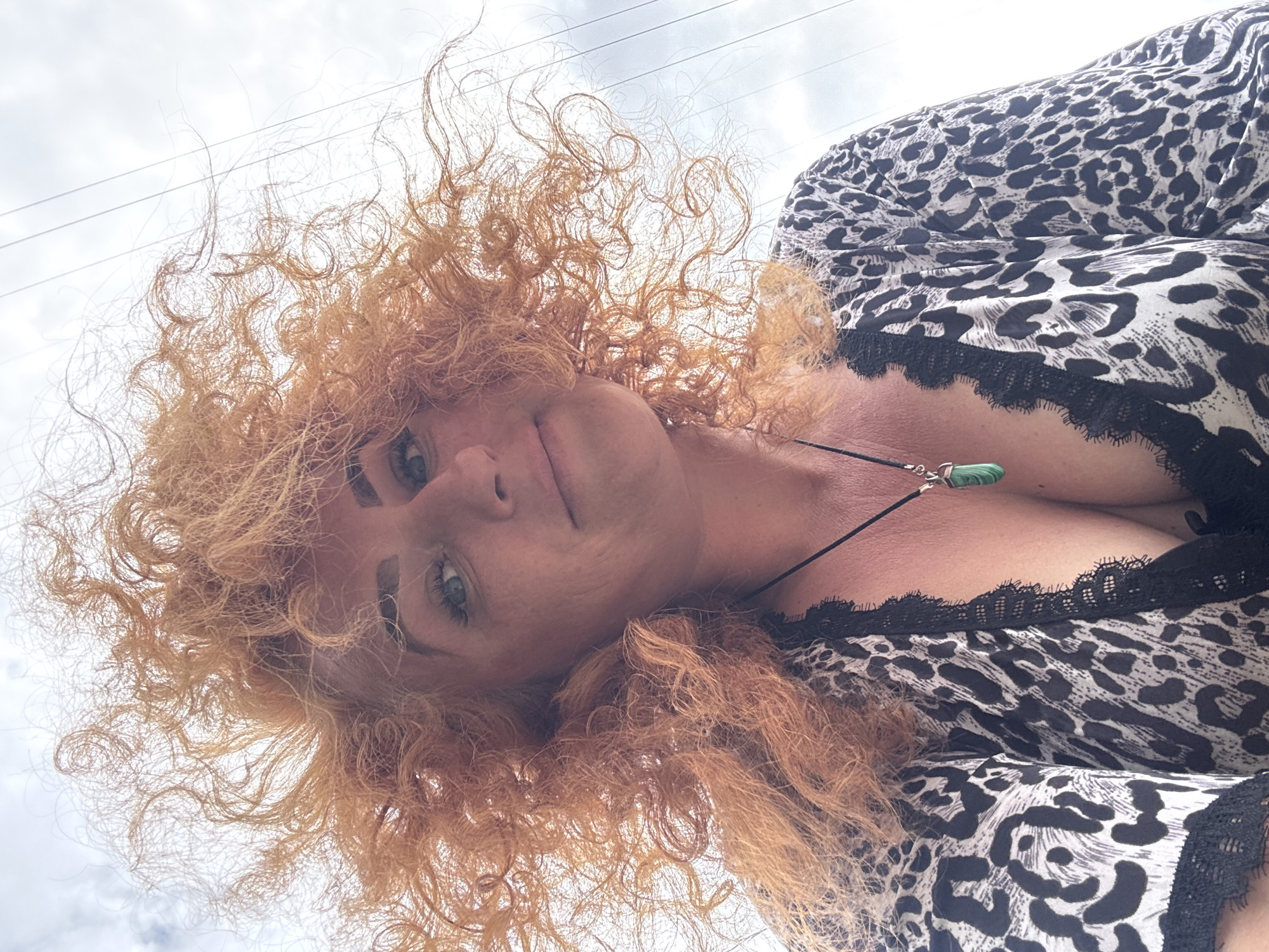 The image shows a close-up, low-angle selfie of a woman with bright orange, curly hair and blue eyes, against a cloudy sky with power lines visible. She is wearing a black and white leopard-print top with black lace trim, and a pendant necklace with a green stone. The setting appears to be outdoors, possibly in Chaikovsky, Russia, though no specific city landmarks are identifiable. The woman is looking up at the camera, with a slight smile. The image is taken from a perspective directly below the woman's face, emphasizing her hair and the sky.