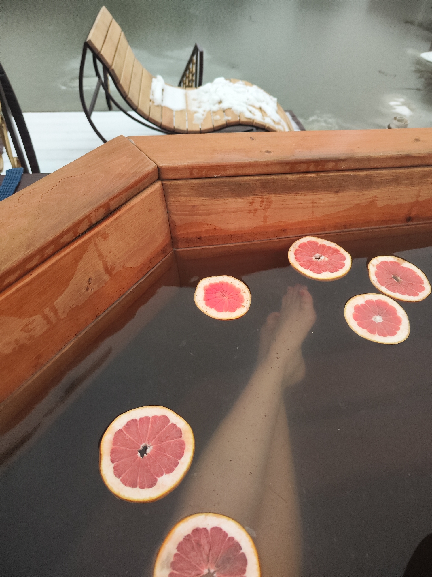 A close-up shot shows a person relaxing in a wooden hot tub, with slices of grapefruit floating on the dark water. The person's legs are submerged. Outside the tub, a snowy scene is visible with a wooden chair. The picture was taken near the city of Chaikovsky, Russia.