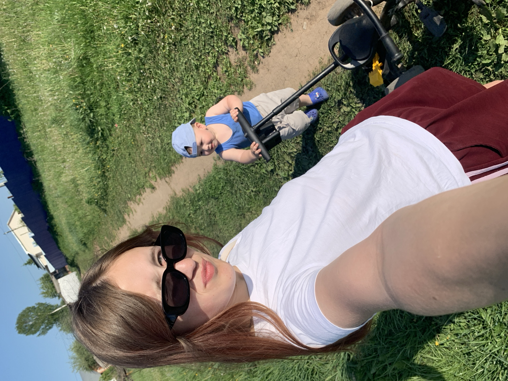 A selfie taken outdoors in Фоки, Russia shows a woman wearing sunglasses and a white t-shirt with maroon pants laying on the grass. Behind her, a small child wearing a blue baseball cap, blue tank top and khaki shorts sits on a walking toy with blue shoes. A black stroller sits off to the side. A house is visible in the background.