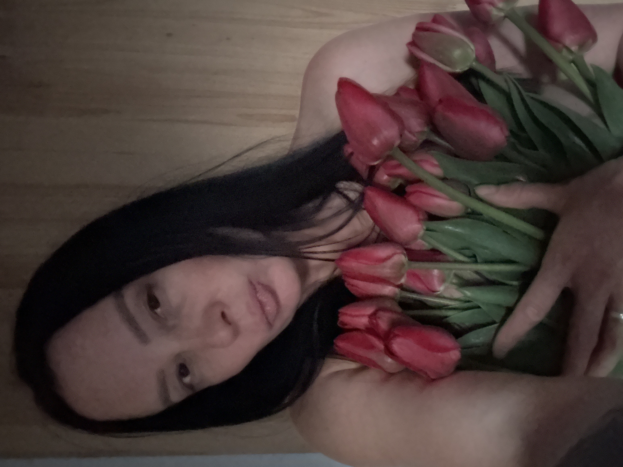 A woman with dark hair is lying on a wooden surface, possibly a floor, holding a bouquet of red tulips. She is looking directly at the camera. Her right hand is visible amongst the tulips, with a ring on one finger. The exact location in Chaikovsky, Russia cannot be determined from this image.