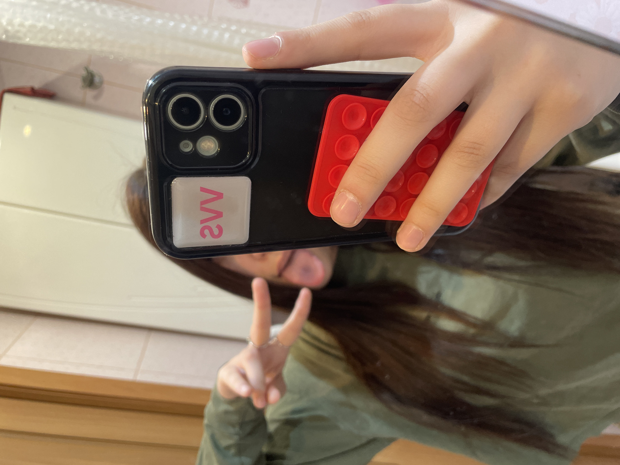 A young person, likely female, takes a mirror selfie in what appears to be an indoor setting, possibly a bathroom or hallway. The individual's face is largely obscured and blurred by the phone and reflection, but she is seen making a peace sign with her left hand, and her tongue is slightly visible, suggesting a playful demeanor. She has long, dark brown hair and is wearing a green long-sleeved top or jacket.
She holds a black smartphone, which features a dual-camera setup, in a black case. Attached to the back of the phone case is a red, rectangular fidget toy with multiple circular indentations, resembling a "Pop It" toy. Additionally, a white square sticker with the reversed pink text "2VV" is visible on the lower left side of the phone's back. The person's right hand, with manicured but chipped nails, is also seen holding the phone.
The background reflected in the mirror includes a large white cabinet or wall surface, a section of light-colored tiled floor or lower wall, and a wooden ledge or counter below. The lighting suggests an indoor setting, likely during the day or in a well-lit room. The overall scene is casual. The image context indicates the location is Saint Petersburg, Russia.