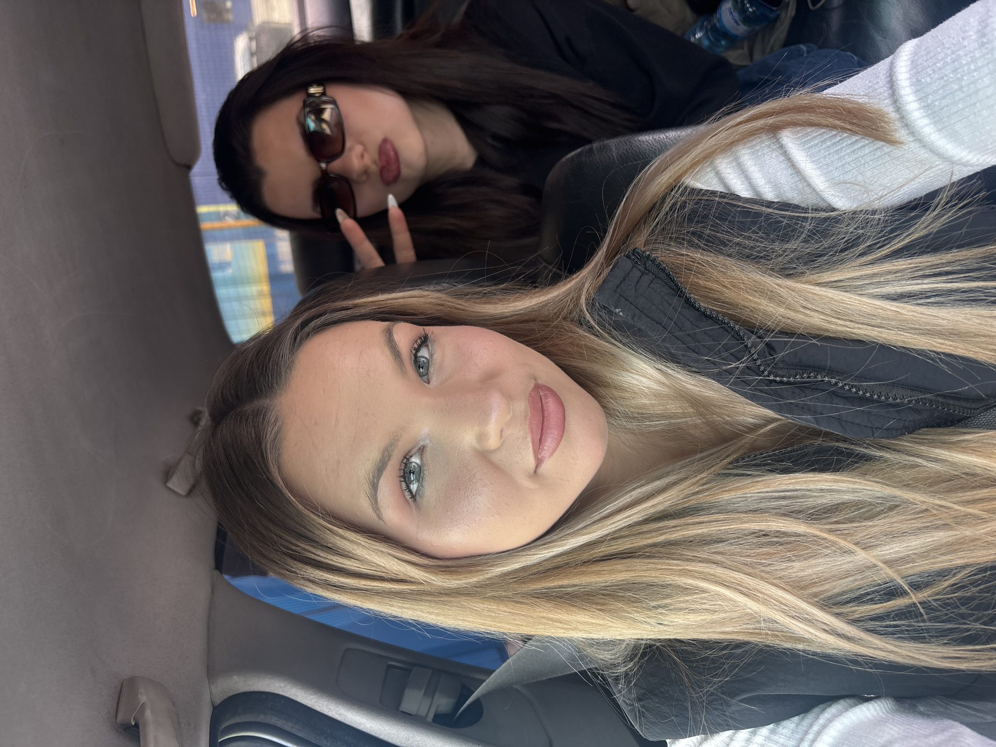 Two women are inside a vehicle. The woman in the foreground has blonde hair and is wearing a black vest over a white sweater. The woman in the background has dark hair and is wearing sunglasses and dark clothing. She is holding up two fingers in a peace sign. The inside of the vehicle is visible. The location is not identifiable as Plovdiv, Bulgaria.