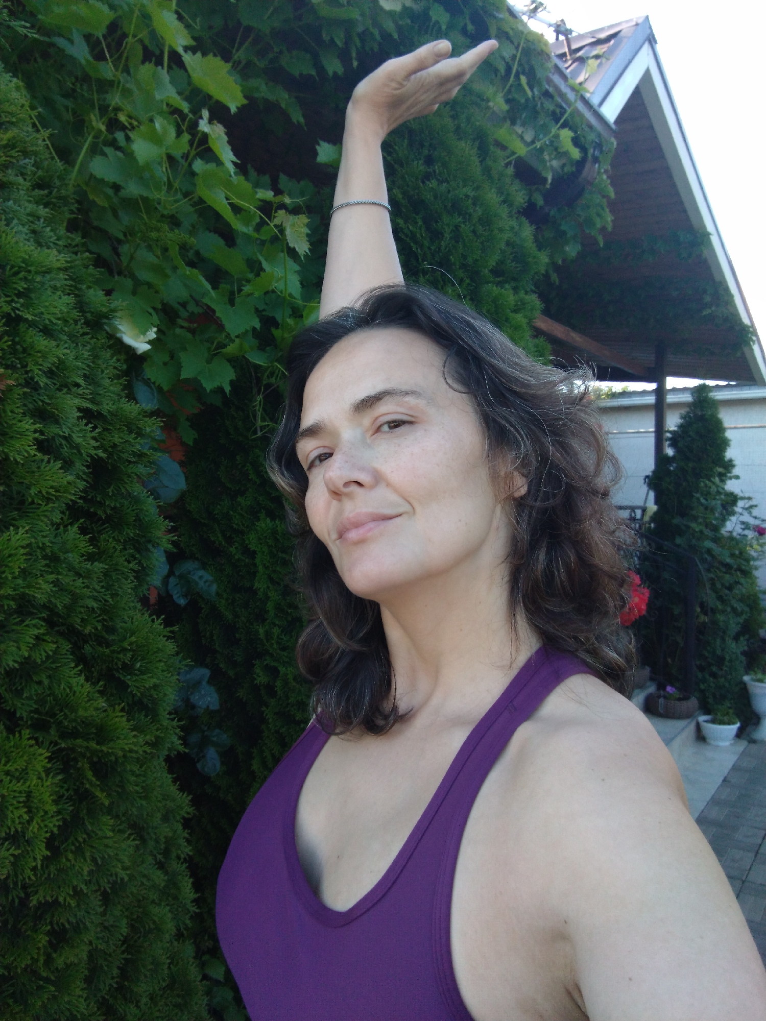 A woman with dark, wavy hair and fair skin is captured in a medium shot, outdoors. She is looking towards the camera, with her left arm extended upwards, fingers slightly splayed. She is wearing a purple tank top. Behind her, dense green foliage and what appears to be a portion of a structure with a sloped roof are visible. The setting suggests a residential garden or yard.