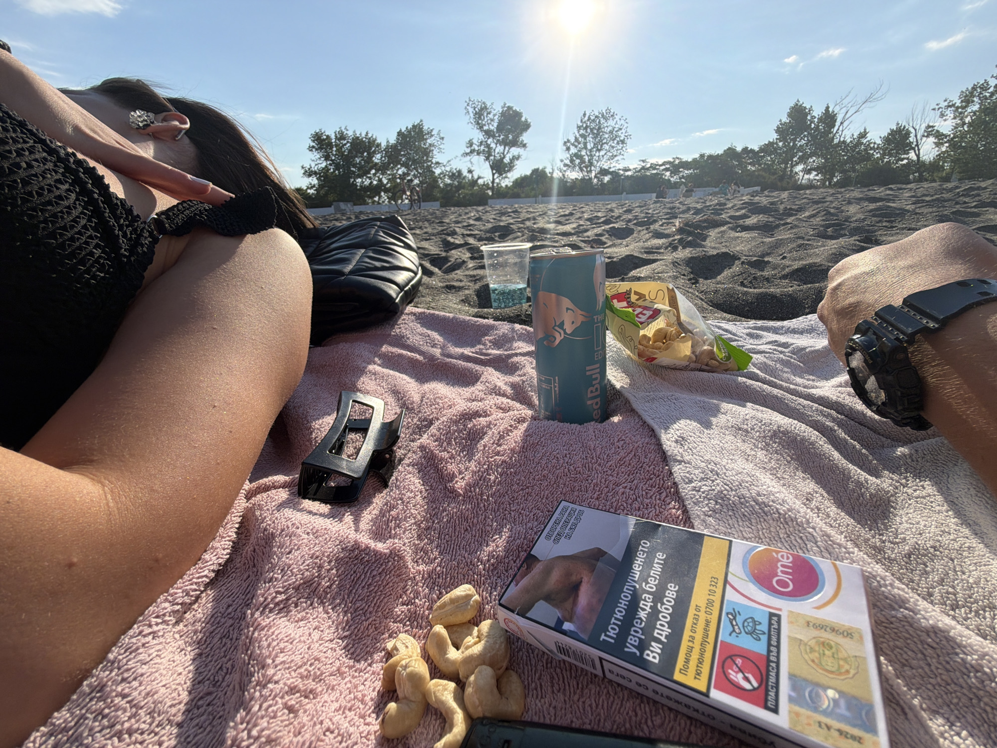 The image shows a close-up of two people relaxing on a beach with black sand, likely in Burgas, Bulgaria, given the context. The sun is shining brightly in a clear blue sky with some scattered clouds.
In the foreground, a pink towel is spread out on the sand. On the towel, several objects are arranged:
* A pack of "Ome" brand cigarettes, featuring a health warning in Cyrillic script.
* A pile of cashew nuts.
* A black hair clip.
* A blue can of Red Bull energy drink, with the iconic bull logo visible.
* A clear plastic cup with a blue liquid and ice.
* A bag of snacks, possibly chips, is partially visible.
To the left, a person's arm and shoulder are visible, wearing a black, textured top with ruffled details. They are lying down, with their hand resting near their face, wearing a large, ornate ring on one finger.
To the right, another person's arm is visible, wearing a black digital wristwatch. Their hand is resting on the towel.
In the background, the beach stretches out, with more sand and some people sitting or walking in the distance. Trees and vegetation line the edge of the beach, and a bicycle is visible further back. The overall scene depicts a relaxed day at the beach.