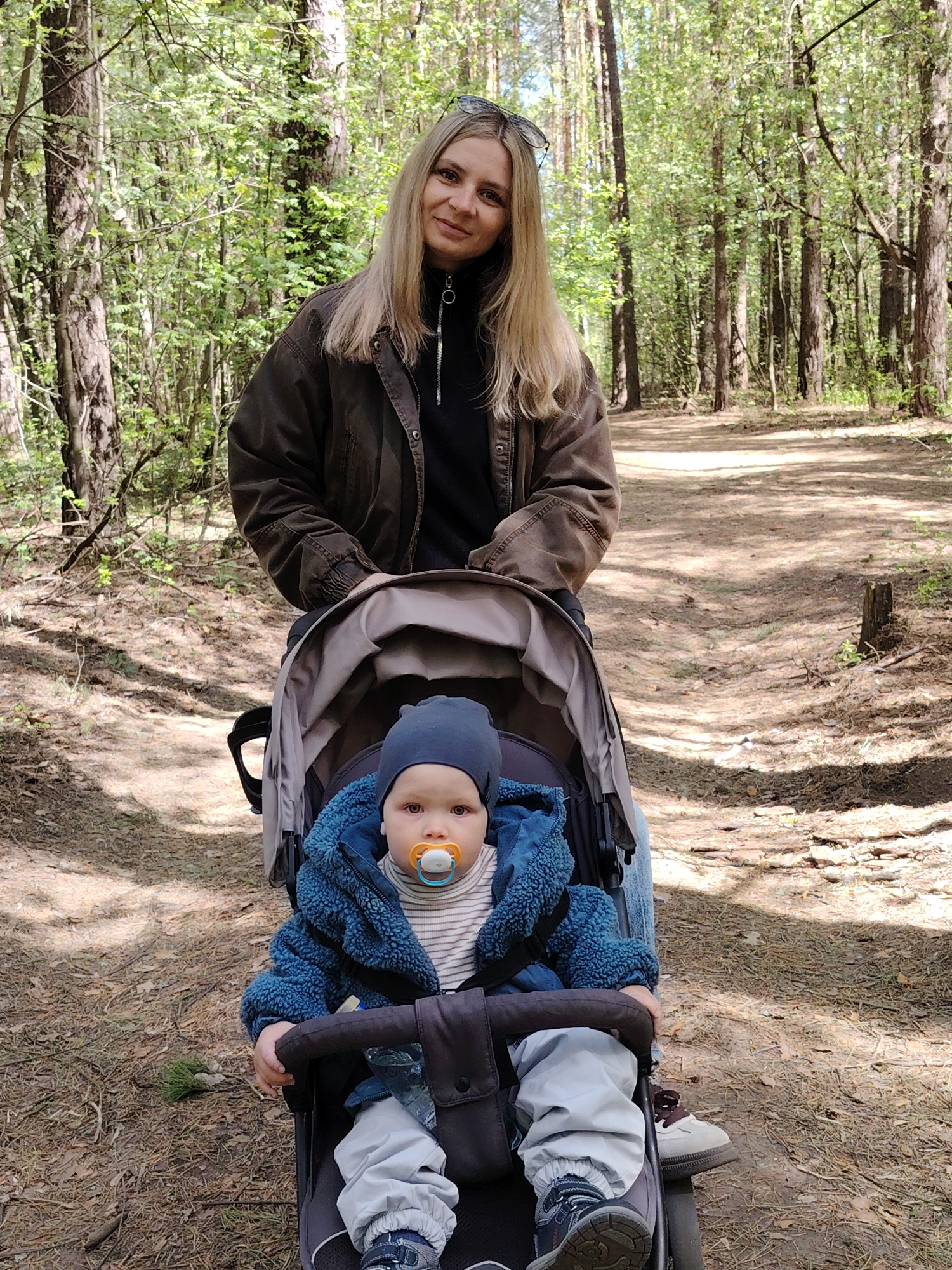 The image shows a woman standing behind a baby stroller on a dirt path in a forest. The woman has blonde hair and is wearing sunglasses on her head, a dark jacket over a black top, and jeans. She is partially visible from the waist up. The stroller contains a baby wearing a navy blue hat and blue jacket, light grey pants, and blue shoes. The baby has a pacifier in its mouth. The stroller has a light gray hood and a dark gray frame. There is a partially visible bottle in the stroller. The background is a dense forest with tall trees and dappled sunlight. The location is the city of Chaikovsky, Russia.