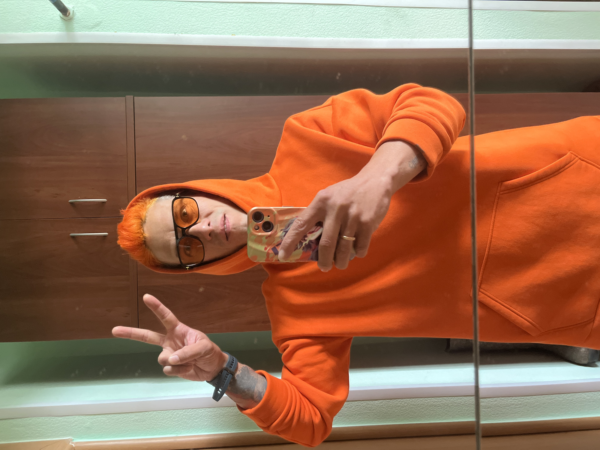 A man is taking a selfie in front of a mirror. He has orange hair and is wearing glasses and an orange hooded sweatshirt. He is making a peace sign with his left hand and holding a phone in his right hand. The background includes a wooden cabinet, a mint green wall, and a ledge. The location appears to be indoors. This is a photograph and nothing indicates the location is the city of Chaikovsky, Russia.