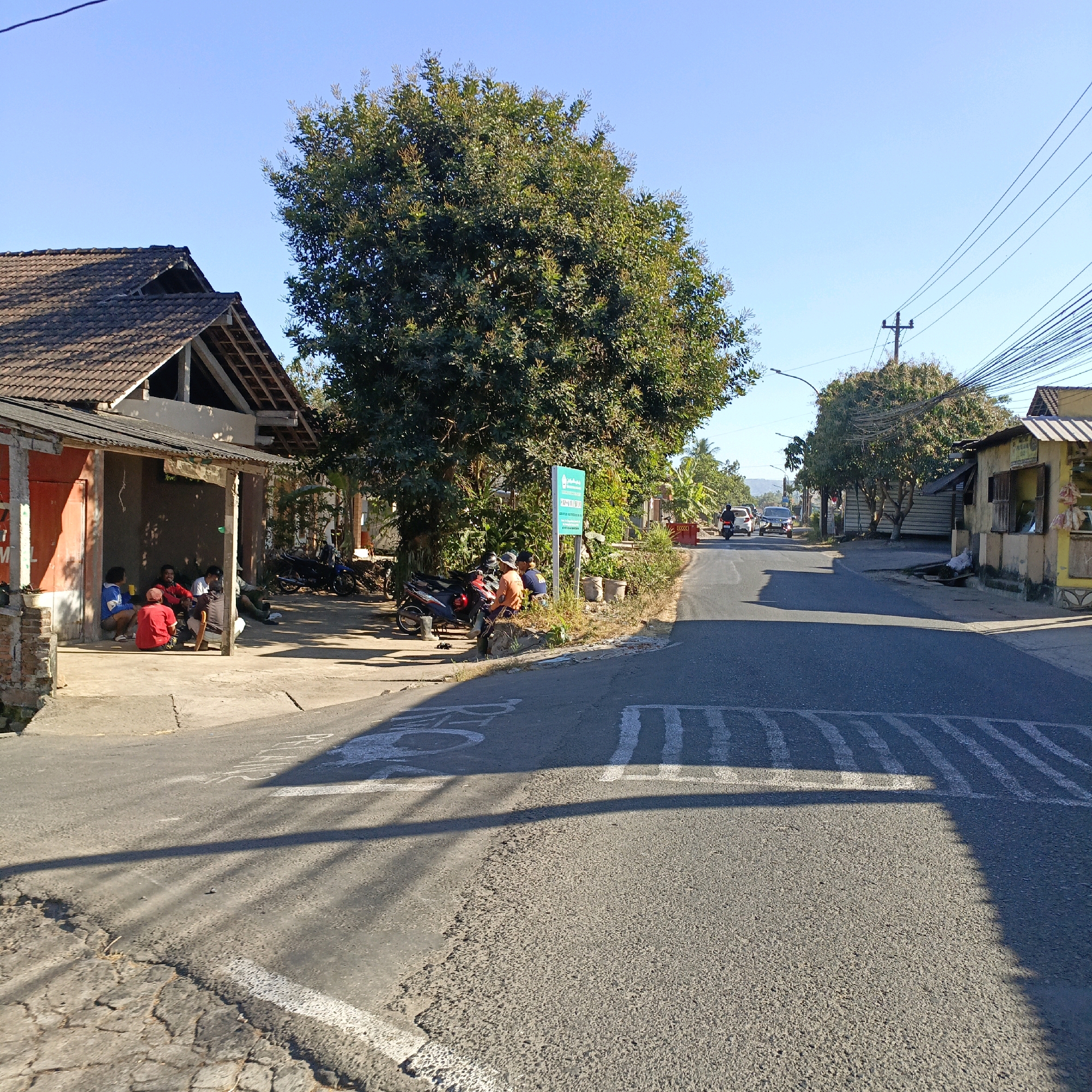The image captures a street scene in Sraten, Indonesia, on a clear, sunny day. In the foreground, the asphalt road shows signs of wear, with a cobblestone section to the left and a painted pedestrian crossing. To the left, a weathered, open-air structure with a tiled roof houses several men sitting on the ground or on low stools. They appear to be engaged in casual conversation, with one man in a red shirt and another in a blue jacket prominent. Motorbikes are parked in the shade of the structure.
Further down the road, a large, leafy tree dominates the center of the frame, casting long shadows across the pavement. A green sign with white text is visible near the tree. On the right side of the road, a small, yellow roadside stall with a striped awning sells goods. Beyond the stall, buildings and more trees line the street, and a power line with multiple cables stretches across the upper right of the image. Vehicles, including a car and motorcycles, are present on the road, suggesting ongoing traffic. The overall environment suggests a rural or semi-urban setting with modest infrastructure.