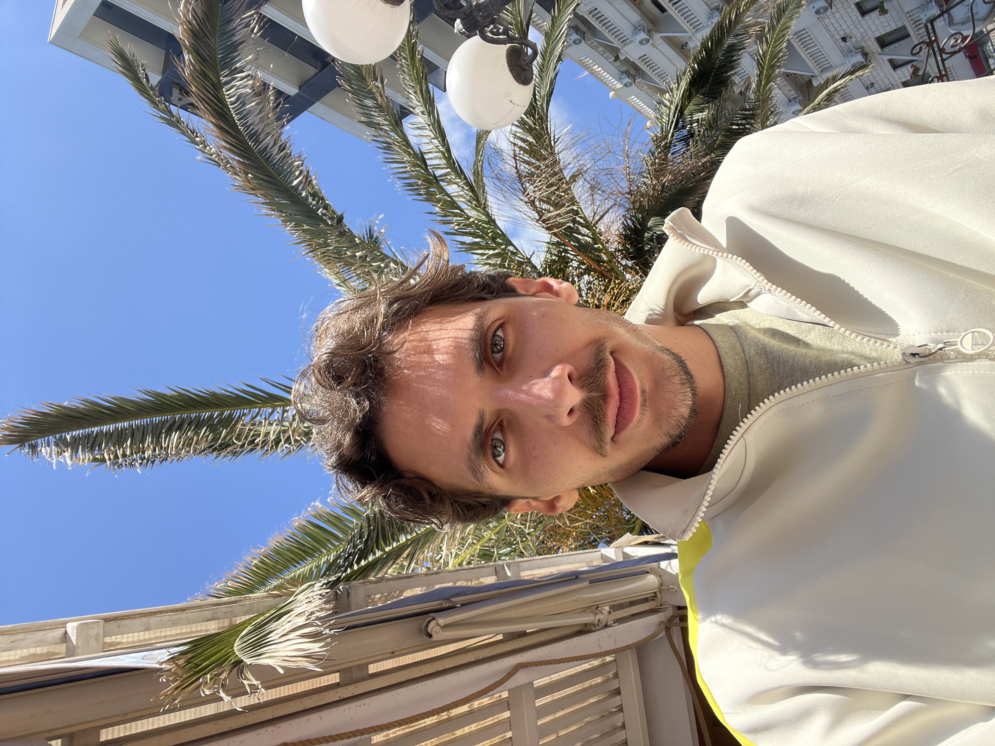 The image is a rotated selfie taken in Budva, Montenegro. The man in the foreground has brown curly hair, green eyes, and a mustache. He is wearing a white jacket with a green shirt underneath. In the background is a palm tree, a white building with lights, and a blue sky. There is a white awning structure with rolled-up shades. The photo appears to have been taken outdoors during the daytime.