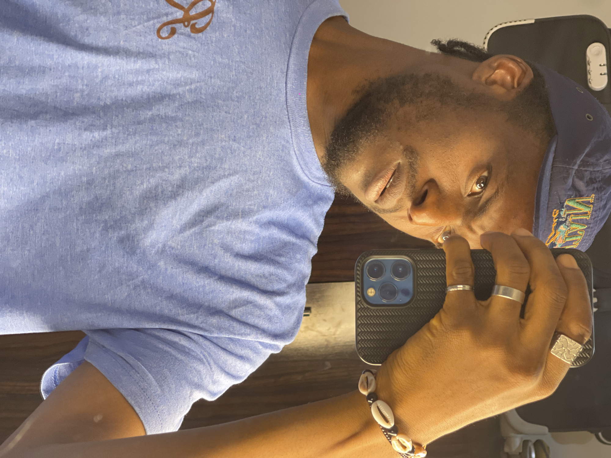 In this close-up selfie, a person with dark skin is holding a phone to take a picture. They are wearing a blue baseball cap and a light blue t-shirt. On their right hand, they are wearing a bracelet with shell beads and several rings. The background appears to be indoors, with a wooden door visible on the right side of the image. There is no identifiable information about the location being in Abuja, Nigeria.