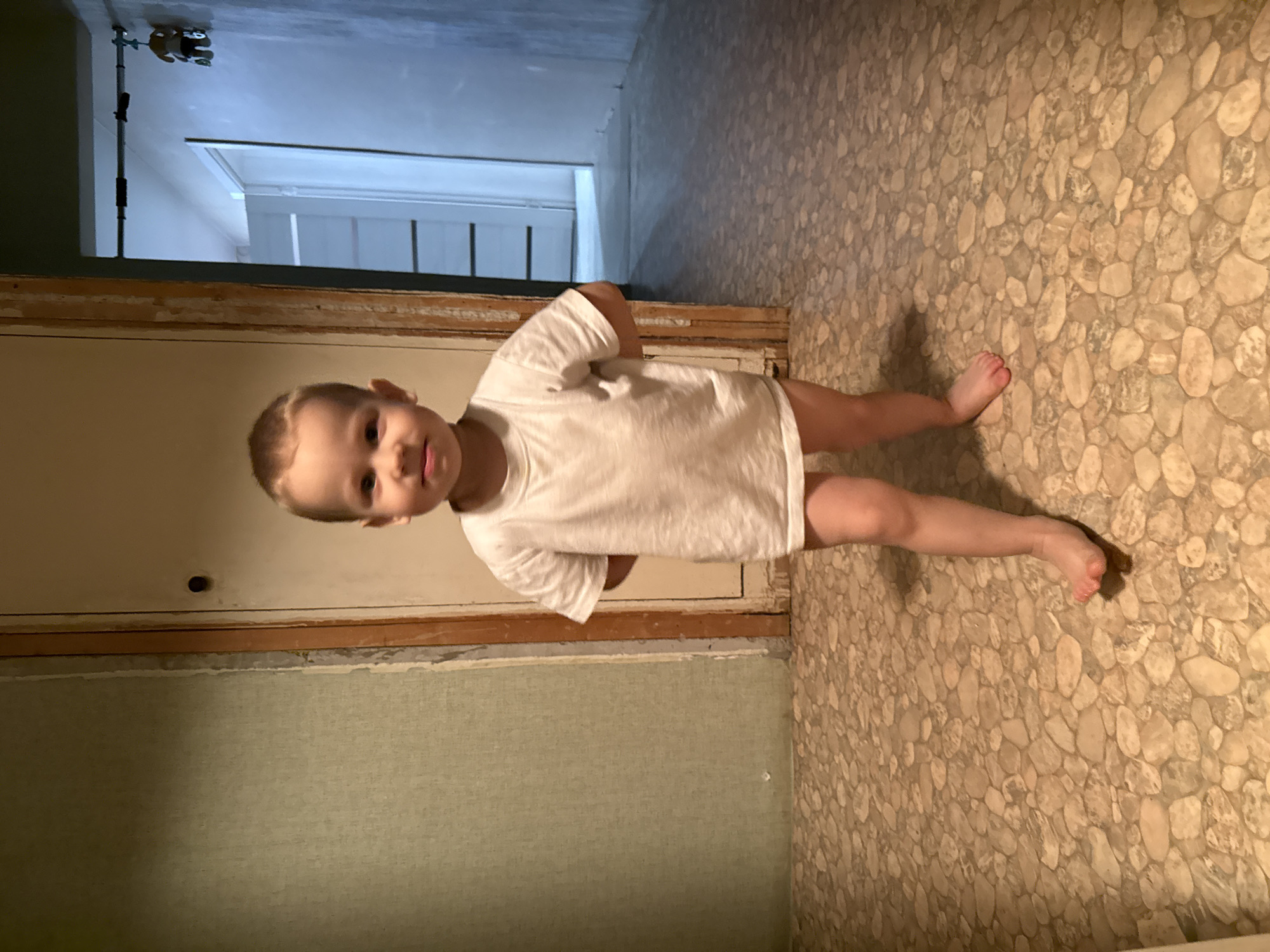 A toddler in a white t-shirt stands barefoot in a room with a stone-patterned floor. The room appears to be under renovation, with unfinished walls. A doorway is visible in the background. There is no information to indicate that the image was taken in Rusanovo, Russia.