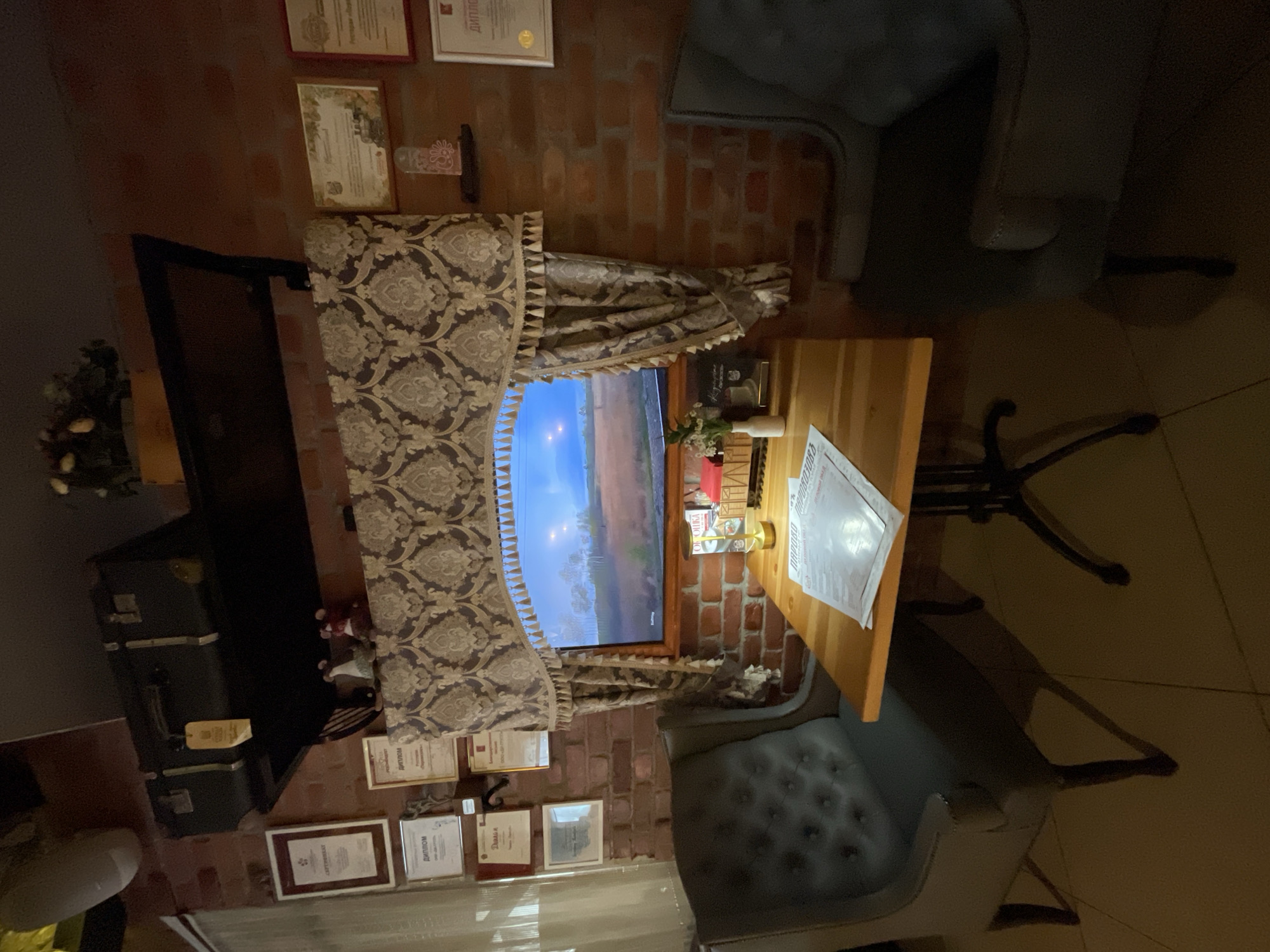 The image shows an indoor scene, possibly a cafe or lounge, with a brick wall as a prominent feature. The room is furnished with a wooden table, two upholstered chairs, and a large trunk. On the wall, several framed certificates and diplomas are displayed. A window looks out onto a landscape, suggesting it might be daytime or early evening. A decorative curtain with a damask pattern hangs above the window.
The scene appears to be set in a relaxed atmosphere. There are no people visible in the image, but the arrangement of furniture suggests a place where people might gather. The presence of framed certificates might indicate an establishment that recognizes achievements or a historical building. The trunk adds a touch of vintage decor. The lighting is dim, casting shadows and highlighting the textures of the brickwork and upholstery.
Visible text includes Cyrillic inscriptions on the framed documents, which appear to be certificates or diplomas. One document on the table also has Cyrillic text, likely a menu or information sheet. The overall impression is of a cozy and perhaps historic interior, with a blend of old and new elements. The location context of Vologda, Russia, adds a specific cultural and geographical element to the interpretation of the scene.