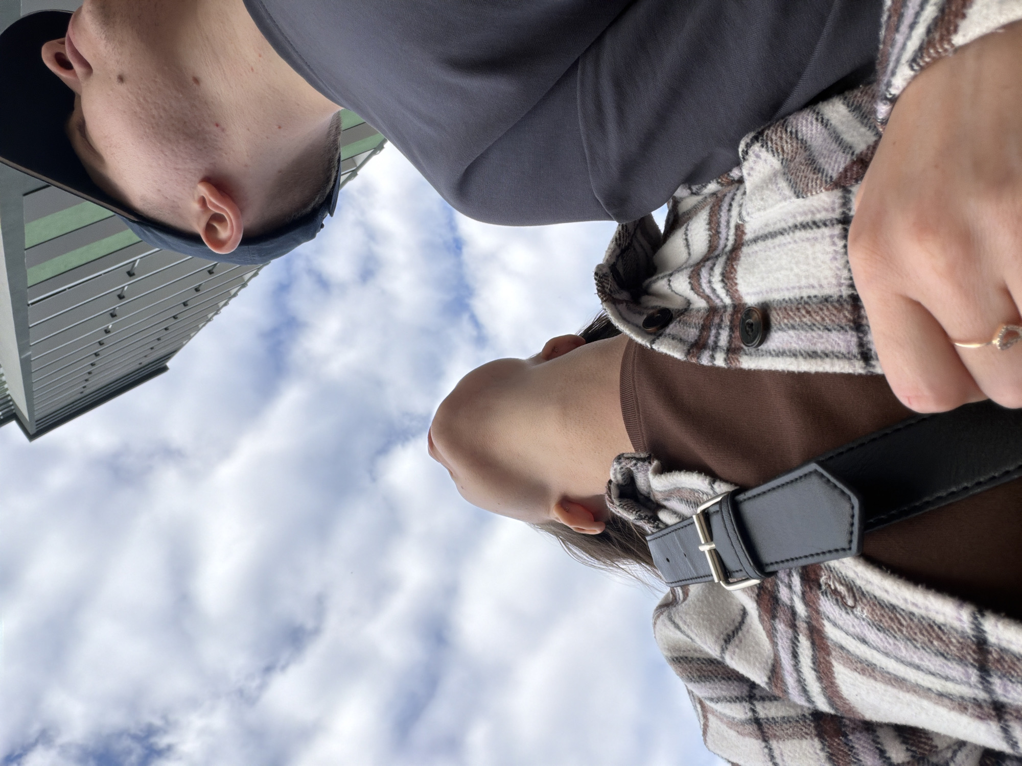 The image shows a low-angle perspective of two individuals against a cloudy sky with a building in the background. The person in the foreground is a woman wearing a plaid shirt over a brown top and a black crossbody bag with a silver buckle. She is looking upwards. To her right and slightly behind her is a man wearing a dark baseball cap and a dark grey t-shirt. He is also looking upwards. The building appears to be modern, with vertical elements. The overall scene suggests an outdoor urban environment in Kyiv, Ukraine, possibly on a slightly overcast day.