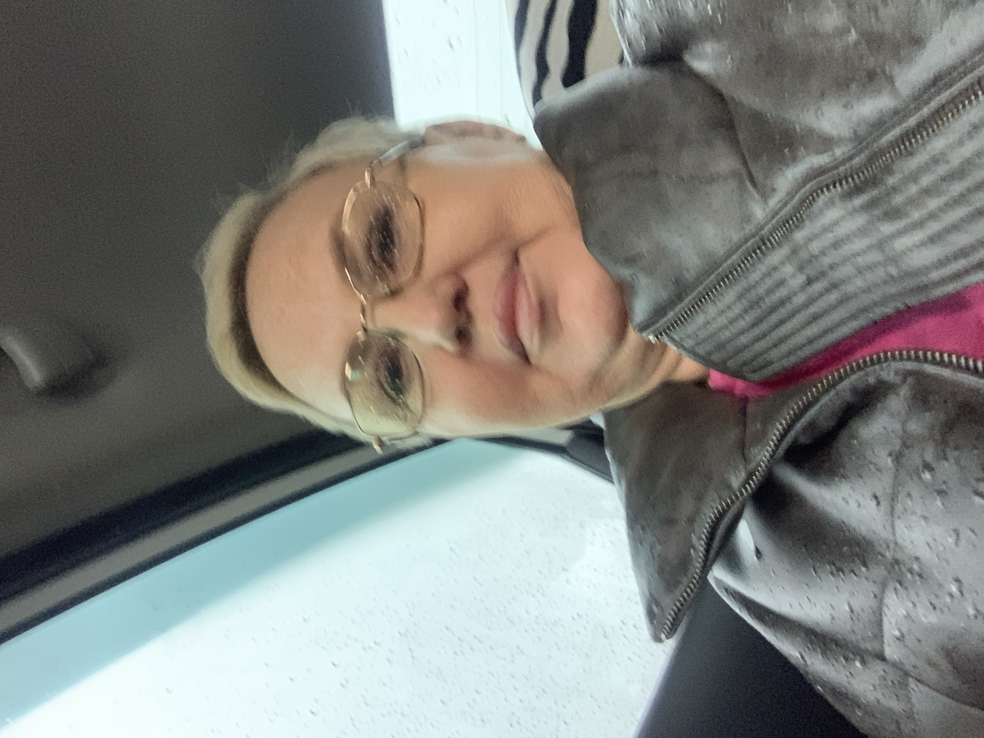 A woman with short blonde hair is wearing glasses and a gray jacket, possibly made of leather, over a pink shirt. The jacket has a zipper. The woman appears to be indoors, possibly in a car, as there is a window with rain droplets visible. There is a handle of a car visible as well. The location in the city of Chaikovsky, Russia is not directly verifiable from the image.