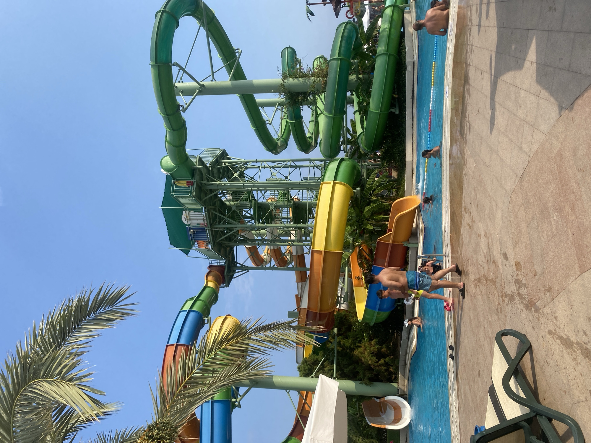 The image depicts a water park located in Aksu, Turkey. Several colorful water slides dominate the scene, with a tall green spiral slide on the right and a multi-colored slide system on the left. A central tower structure supports some of the slides. The pool area is filled with clear blue water, and several people are visible enjoying the amenities. In the foreground, two men are standing near the pool, one appearing to be a young adult and the other an adult. A lounge chair is positioned in the lower left corner of the frame. The environment is sunny with a clear blue sky and palm trees visible, suggesting a warm climate.