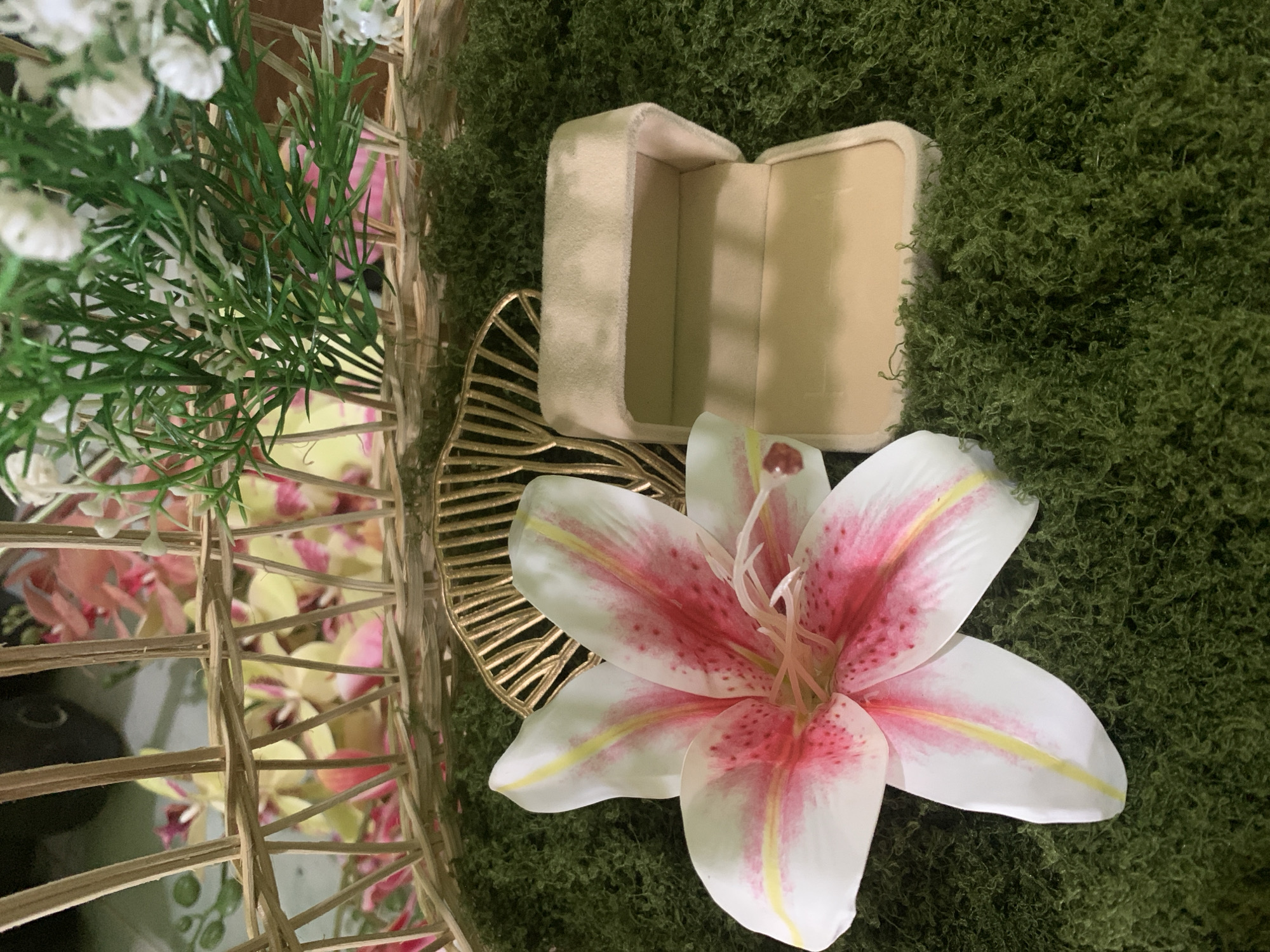 This image depicts a close-up scene of decorative elements arranged on a green, moss-like surface. In the foreground, a single, large, white and pink lily lies to the left. To the right of the lily, an open, light beige jewelry box made of velvet is visible. Behind the lily and jewelry box, a golden, decorative piece with a fan-like design is partially obscured. The background features a woven basket filled with artificial pink and yellow orchids and green foliage. The scene is lit from above, casting soft shadows. No people or actions are depicted. The image does not provide any information about the location in the city of Kuala Pembuang, Indonesia.
