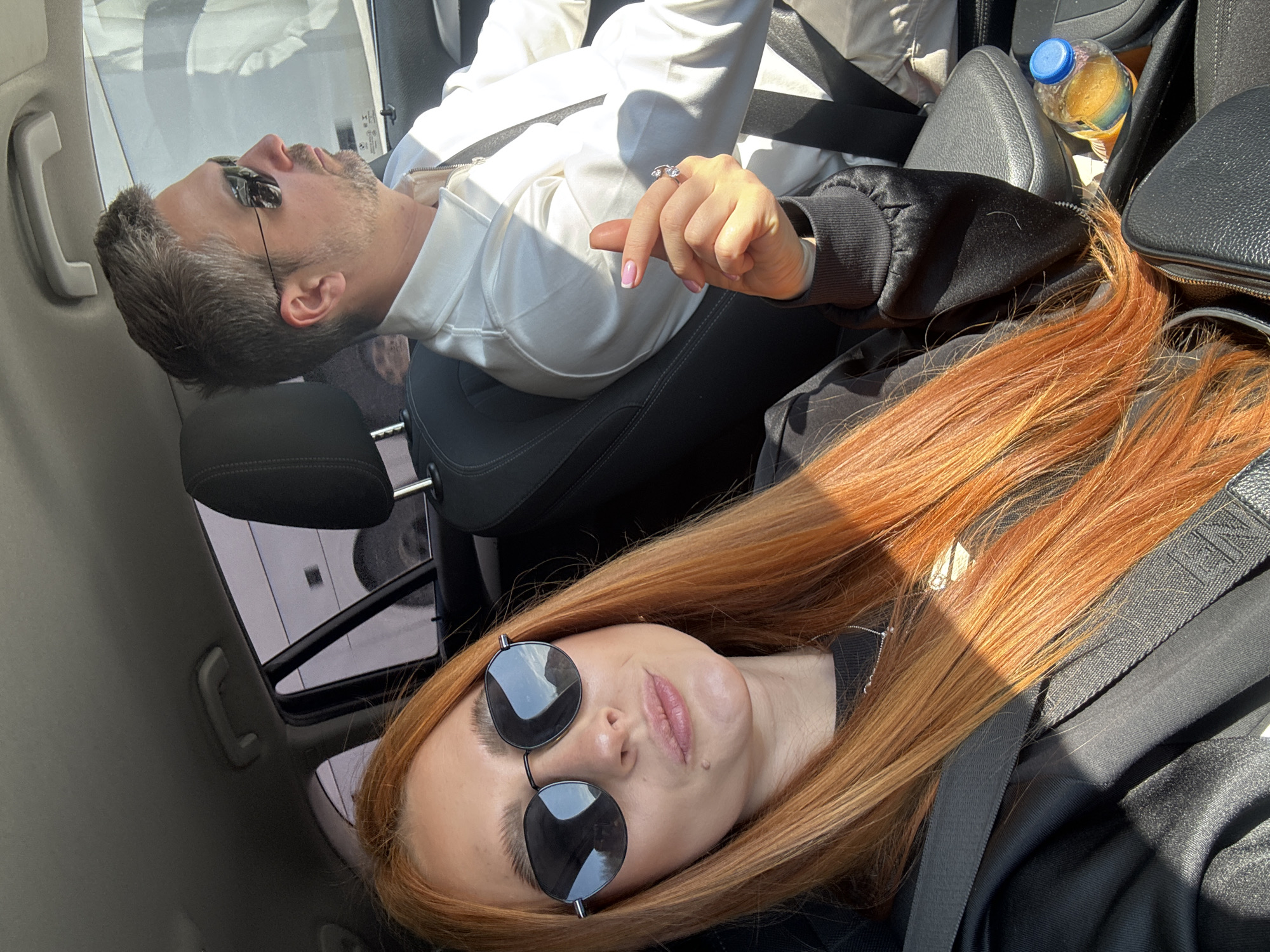 The photo appears to be taken inside a car, possibly in Mišići, Montenegro given the query. Two people are visible. A woman with long auburn hair and wearing round sunglasses is seated in the foreground, presumably in the passenger seat. She is wearing a black top and has a seatbelt on. A man is seated behind her, also wearing a seatbelt. He has grey hair and appears to be wearing eye masks. He is wearing a white shirt or jacket. A water bottle is visible on the car seat next to him. The car has a sunroof. The photo is taken at an angle.