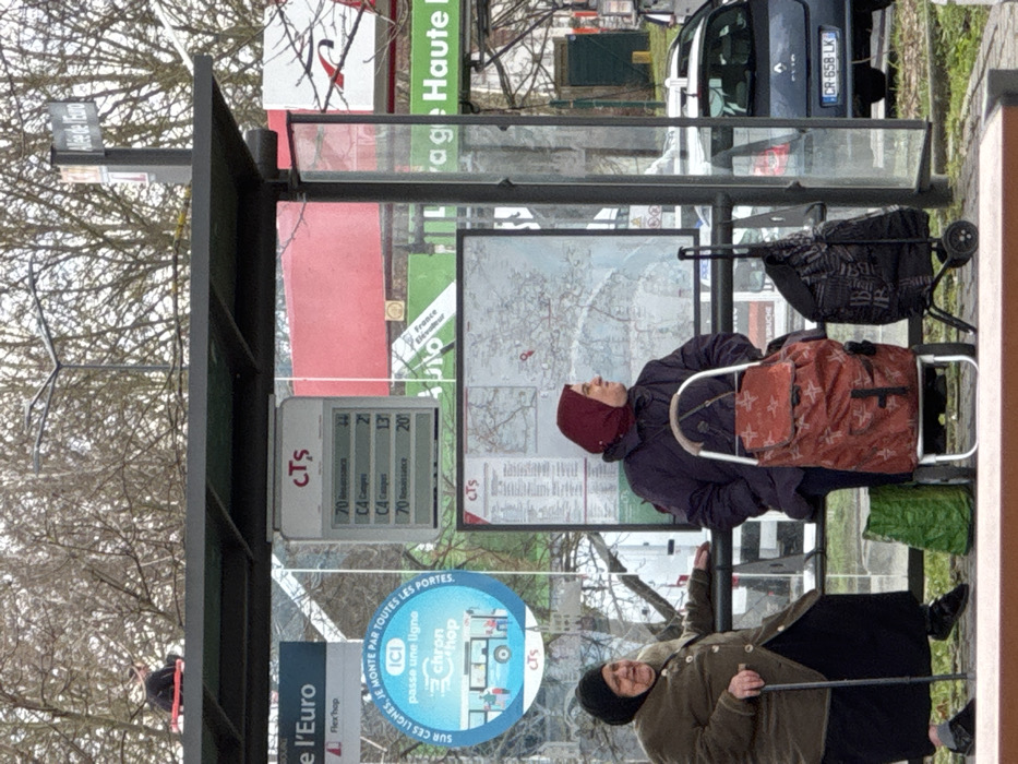 At a bus stop on Allée de l'Euro in Oberhausbergen, France, two women are seated within a transparent bus shelter. The woman on the left, an elderly individual with a black head covering, wears a brown jacket and black skirt, holding a black cane while looking to her left. The woman on the right, wearing a dark purple jacket and a red head covering, is seated in profile, looking towards the right. Next to her are two shopping trolleys: one with an orange and star-patterned bag, and another with a dark bag. A green bag rests on the ground beside the orange trolley.
Above the women, an electronic display board shows "CTS" and bus route information (lines 70 and C4), with a bus network map below it. A blue circular sign promoting "Chron'hop" is visible to the left. A separate sign to the right confirms the location as "Allée de l'Euro".
Above the bus shelter's dark roof, an individual is seated, wearing dark clothing and appearing to have a red object beside them. The background features bare trees, a dark blue Renault Clio car (license plate CR-658-LK), and signage for what appears to be a service station or car wash facility ("Auto," "Lavage Haute Pression").