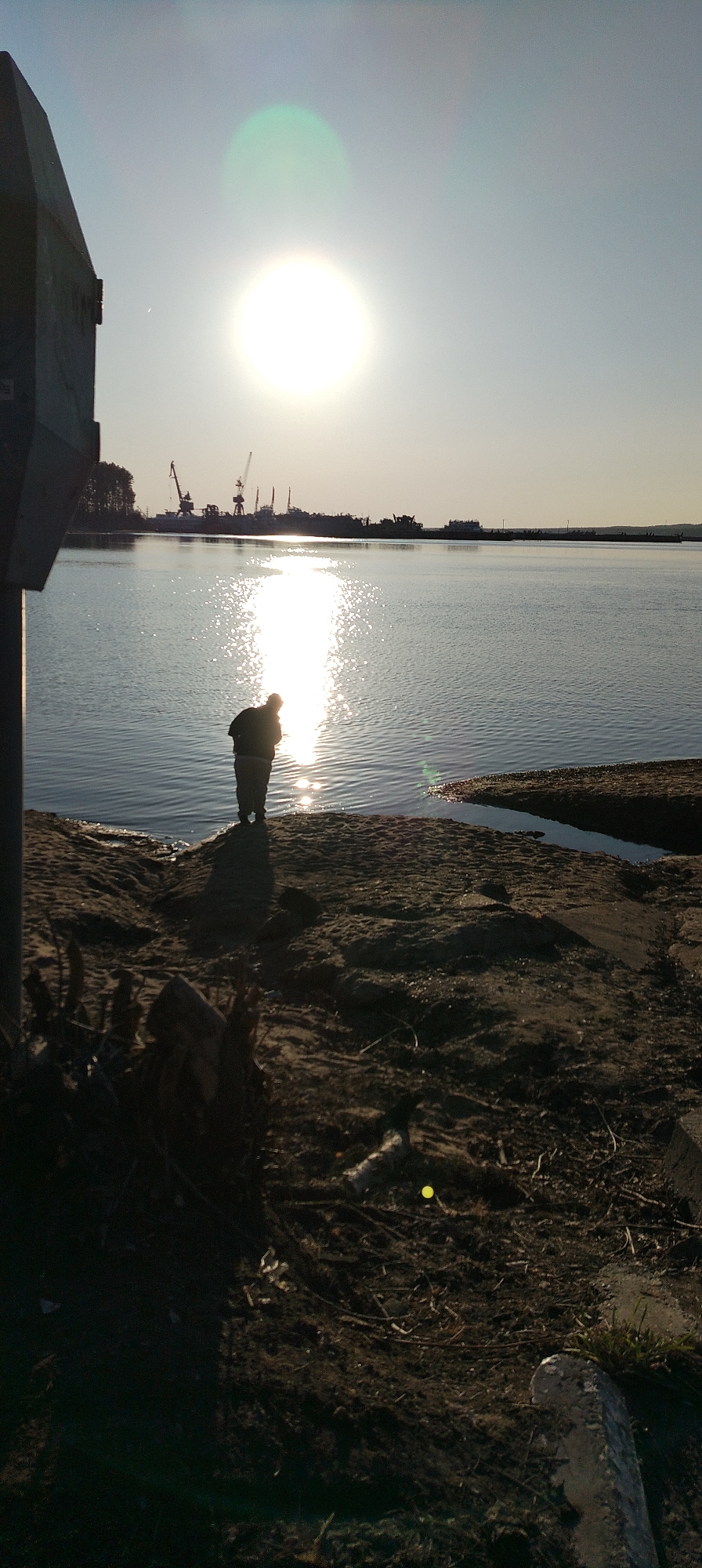 The image depicts an individual standing at the edge of a body of water, possibly a river or bay. The person is facing the water, with their back towards the viewer. The sun is positioned high in the sky, creating a bright glare on the water surface. In the background, there are silhouetted structures that appear to be cranes and industrial facilities, suggesting a port or industrial area. The immediate foreground shows a sandy or muddy bank with some vegetation. A concrete structure or pole is visible on the left side of the frame. The environment suggests a location in an urban or industrial area near a significant body of water.