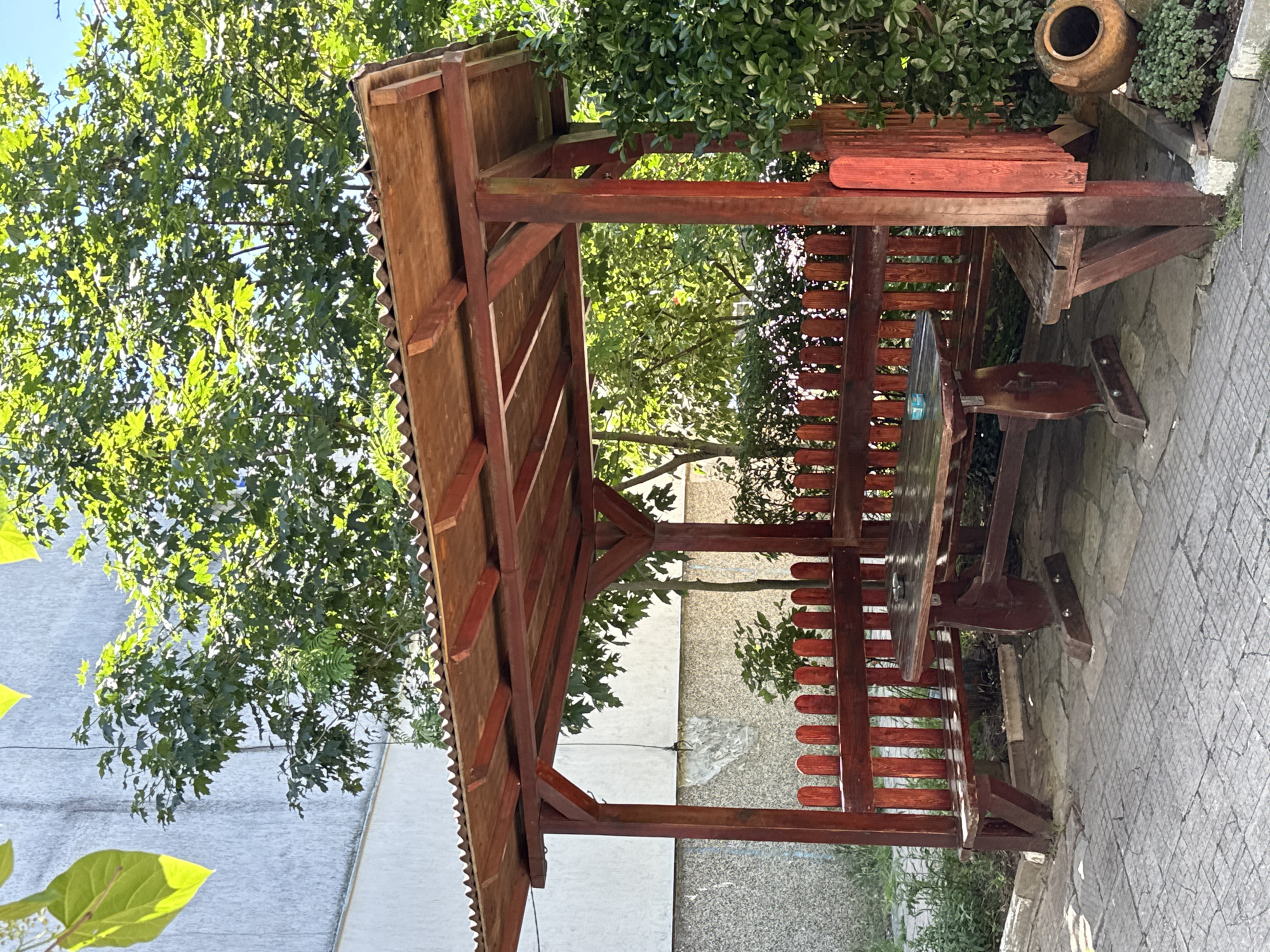The image shows a wooden gazebo with a table and benches situated on a paved outdoor area. The gazebo has a corrugated roof and red-painted wooden posts and railing. A wooden table and benches are placed inside the gazebo. Behind the gazebo, there is lush green foliage and a building. To the right of the gazebo, a clay pot is visible. The scene appears to be in a residential or recreational area. The specific location in Vratsa, Bulgaria, is not determinable from the image.