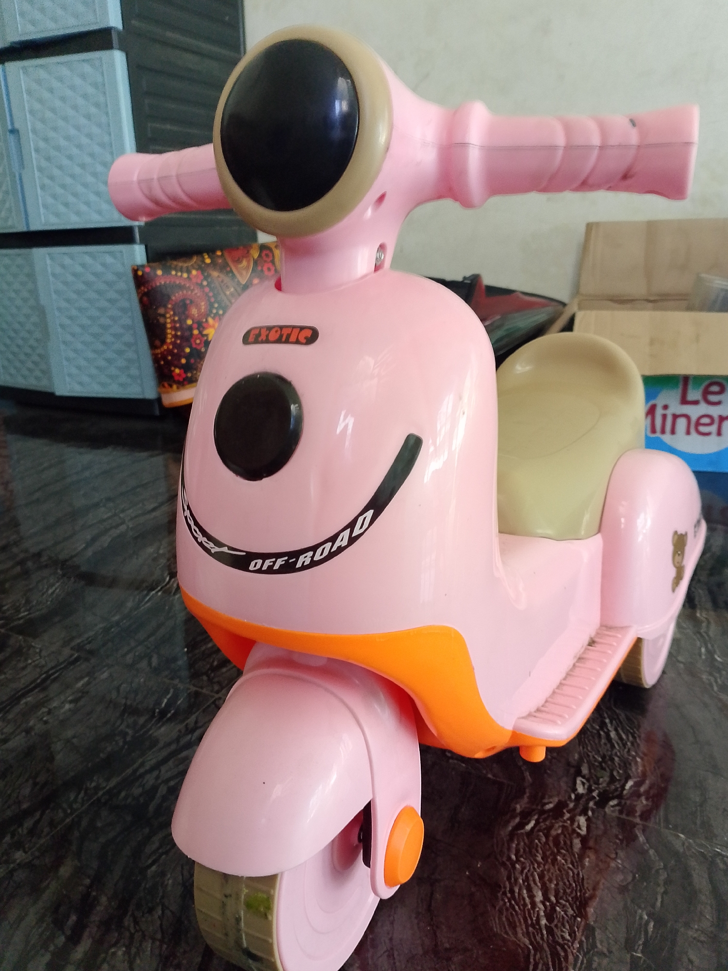 A close-up, eye-level, medium shot captures a pink toy scooter with an orange bottom trim. The scooter is stationary on a dark, reflective surface, possibly a table. The front of the scooter features a large black circular headlight with a beige rim, and two pink handlebars extend outwards. A black decal with the word "EXOTIC" is affixed to the front body of the scooter, above a black circular emblem. Below this, a curved black decal reads "OFF-ROAD". A light-colored seat is visible in the middle of the scooter. To the right of the seat, a small brown teddy bear decal is present on the rear fender. In the background, out of focus, are blue plastic storage drawers to the left, and cardboard boxes and a blue and white bottle to the right. The overall lighting is bright, highlighting the toy. The image was taken in Situwangi, Indonesia.