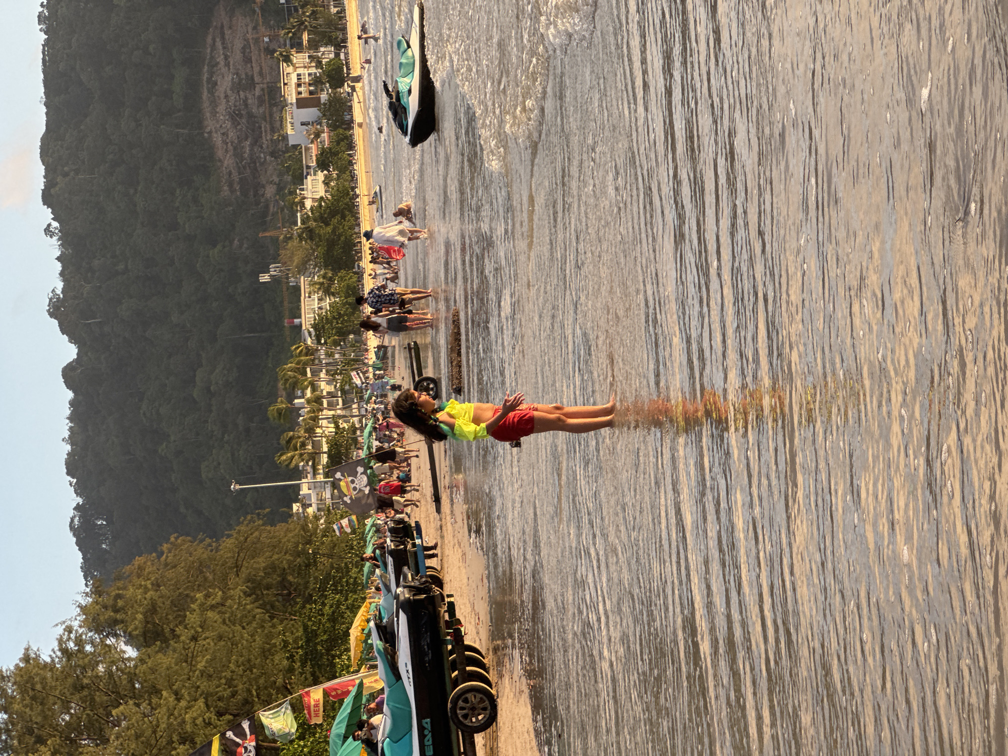 The image depicts a beach scene in Patong, Thailand, during what appears to be late afternoon or early evening, as indicated by the warm lighting. In the foreground, a young girl stands in shallow water, wearing a bright yellow and green top and red shorts. Her reflection is visible on the wet sand. Behind her, a line of jet skis on trailers is parked on the beach, with umbrellas and more people visible further back. The background shows lush green hills, hotels, and some construction. The ocean is calm with gentle waves lapping at the shore. Several people are scattered across the beach and in the water, some walking and others engaging in recreational activities. Pirate-themed flags are visible among the beach amenities.