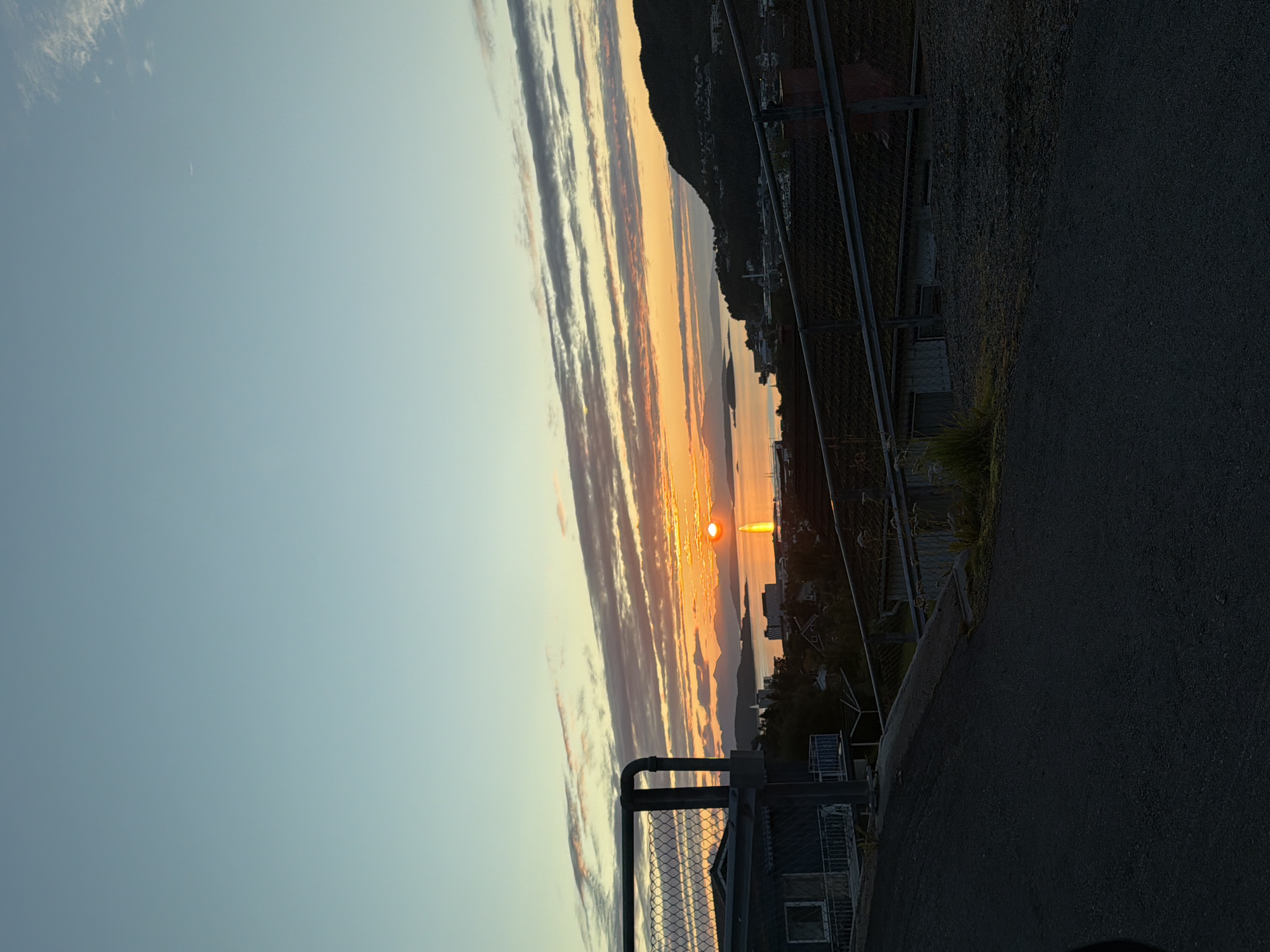 The image shows a sunset over the city of Harstad, Norway. The sun is low on the horizon, casting a warm orange glow across the sky. The sky is a mix of blue and orange, with streaks of clouds. In the foreground, there are houses and a fence. The houses are dark and silhouetted against the sunset. The fence is made of metal and is covered in wire mesh. The image appears to have been taken from a slightly elevated position, possibly a hillside or a road. The overall impression is one of a peaceful and serene evening in a coastal city.