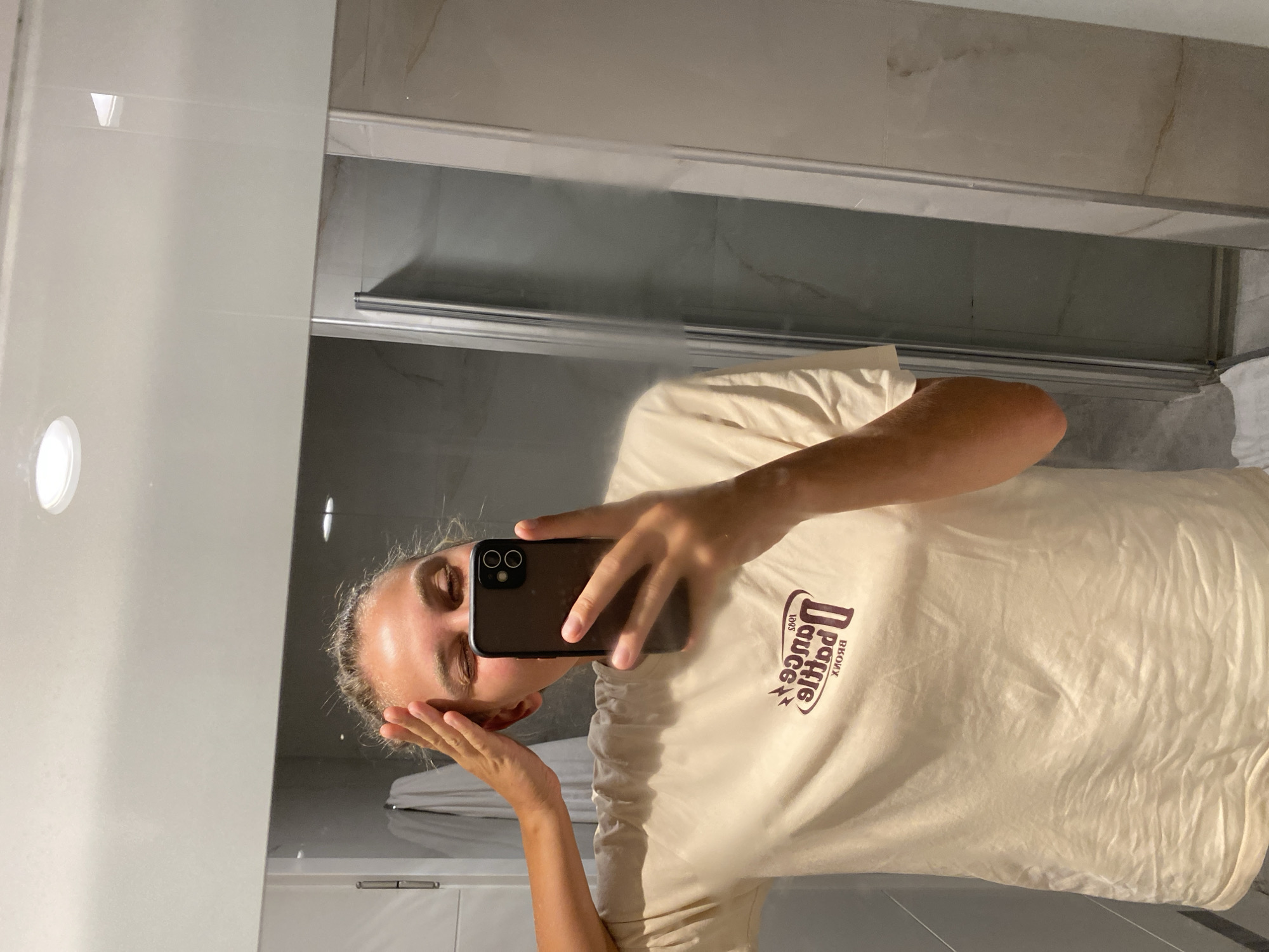 A young woman with blonde hair pulled back is taking a selfie in a bathroom. She is wearing a tan t-shirt with a graphic design on the front. Her right hand is raised to her forehead, and she is winking at the camera. The background shows a tiled wall and a glass shower door. The location is identified as Aksu, Turkey.