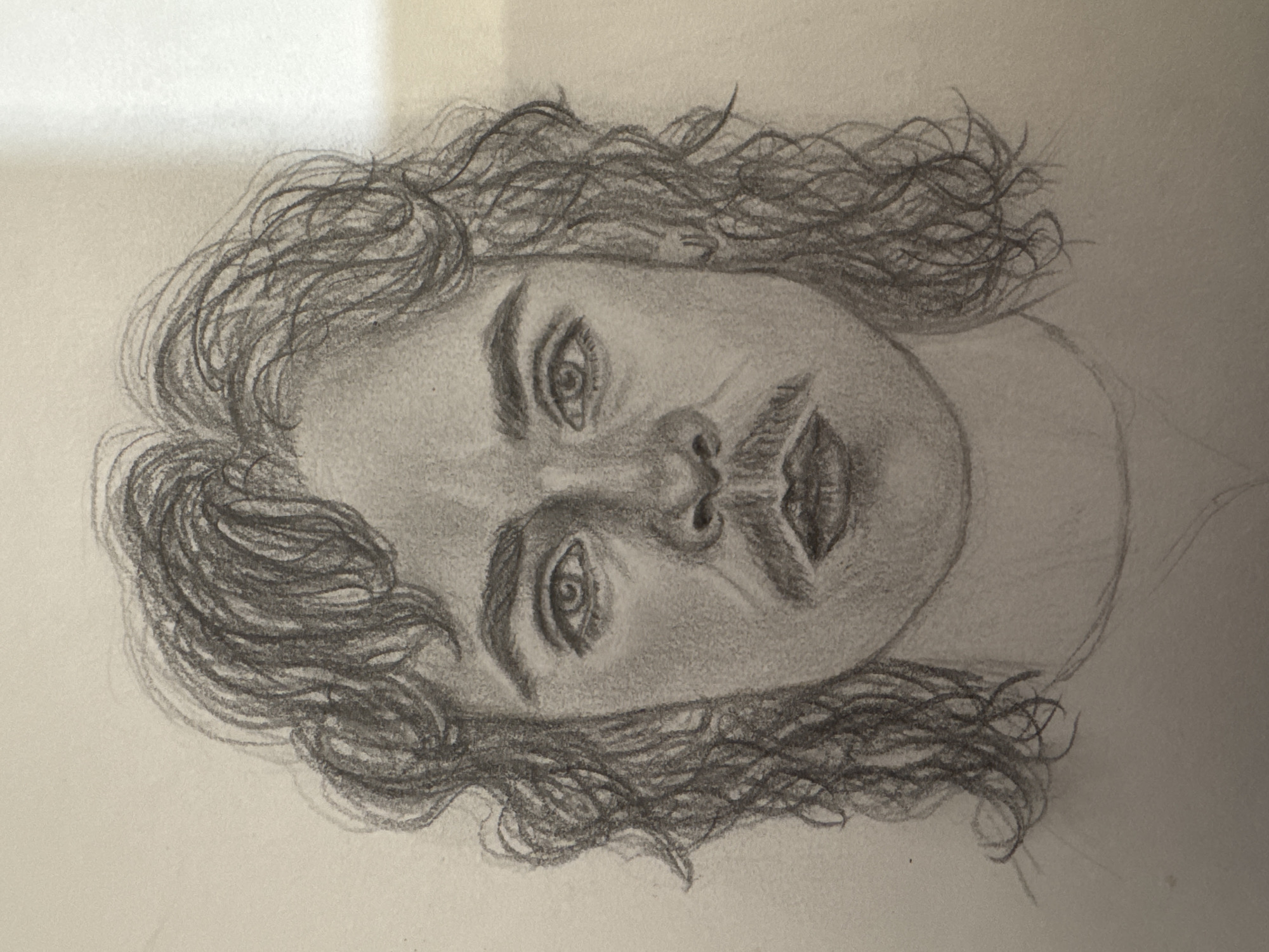 This is a pencil sketch of a young man's face, with curly, dark hair framing his head and extending down to his shoulders. He has a prominent mustache and his lips are slightly parted. The sketch captures a serious or intense expression. The background is a plain, lightly textured surface, suggesting it was drawn on paper. There is no indication of a specific location in Mikhaylovsk, Russia or any other identifiable environment.