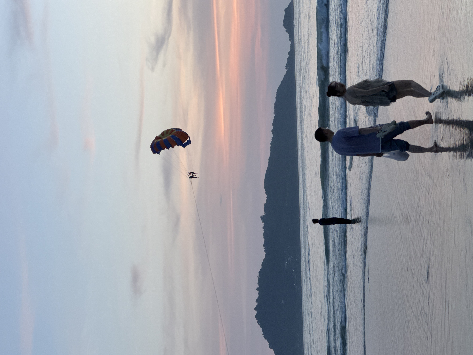 At dusk on Patong Beach, Thailand, the ocean meets the shore under a pastel sky. A lone figure stands in the shallow water, silhouetted against the waves. On the wet sand, a couple walks hand-in-hand away from the viewer, the man carrying a plastic bag and the woman looking back over her shoulder. In the distance, a parasailer with a colorful parachute is suspended in the air above the sea, towed by an unseen boat. The silhouette of a mountainous coastline forms the horizon.