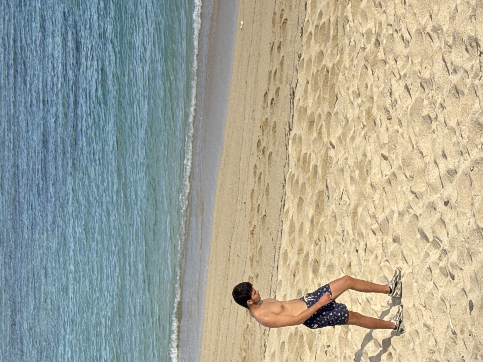 An unidentified shirtless male, wearing dark patterned swim shorts and athletic shoes, walks alone on a sandy beach in Kuta Selatan, Indonesia. The male is positioned in the lower left quadrant of the image, facing towards the left with his head down. The beach is characterized by its light-colored sand with visible footprints. In the background, the ocean displays shades of blue and green with gentle waves lapping the shore. The lighting suggests it is daytime.