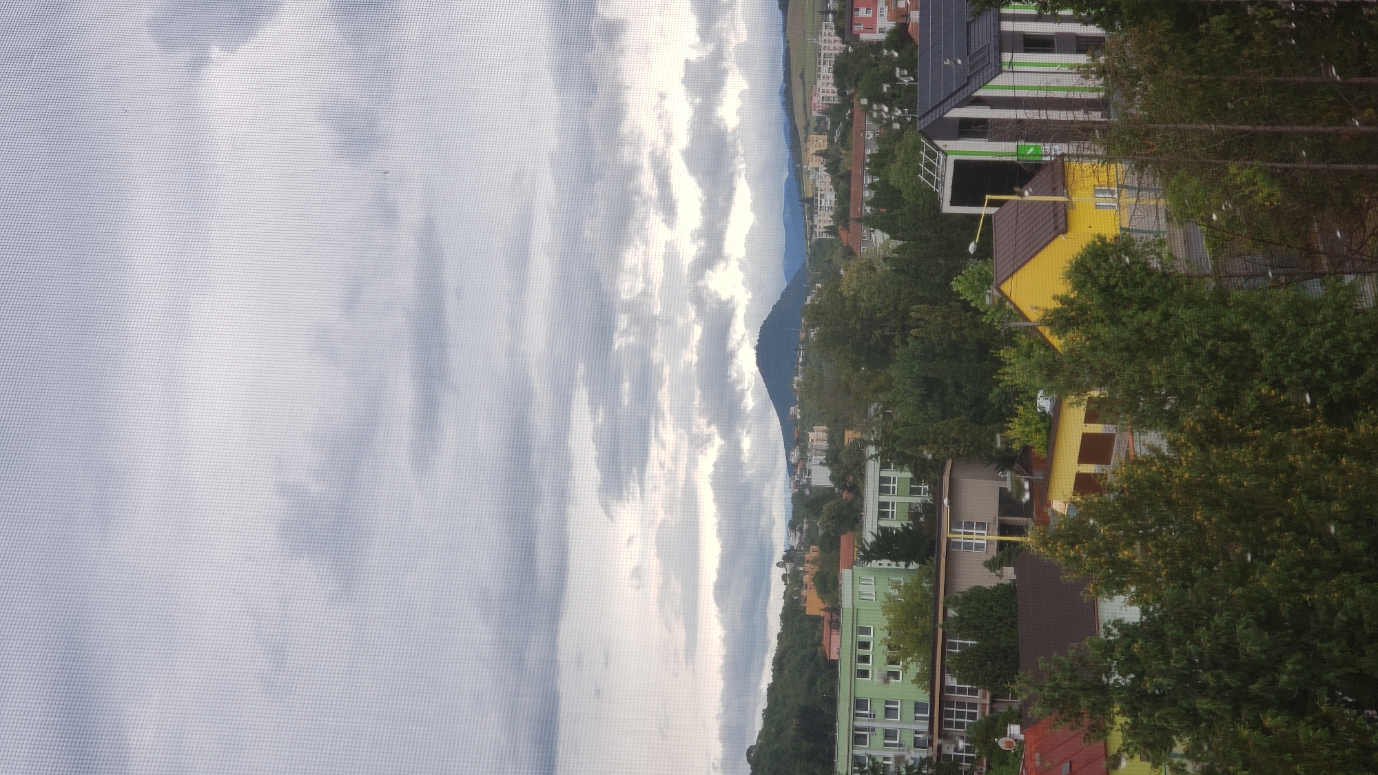 The image shows a view of a city with buildings and trees under a cloudy sky. In the foreground, there are trees and some houses. Behind them, there are larger buildings, including what appears to be apartment blocks. In the distance, there are more buildings, hills, and a prominent conical hill with some structures on top. The sky is filled with heavy, gray clouds, suggesting overcast weather. The image appears to be taken from a window, as evidenced by the slight mesh pattern visible throughout. The setting is a residential area with a mix of older and modern architecture, interspersed with lush greenery.