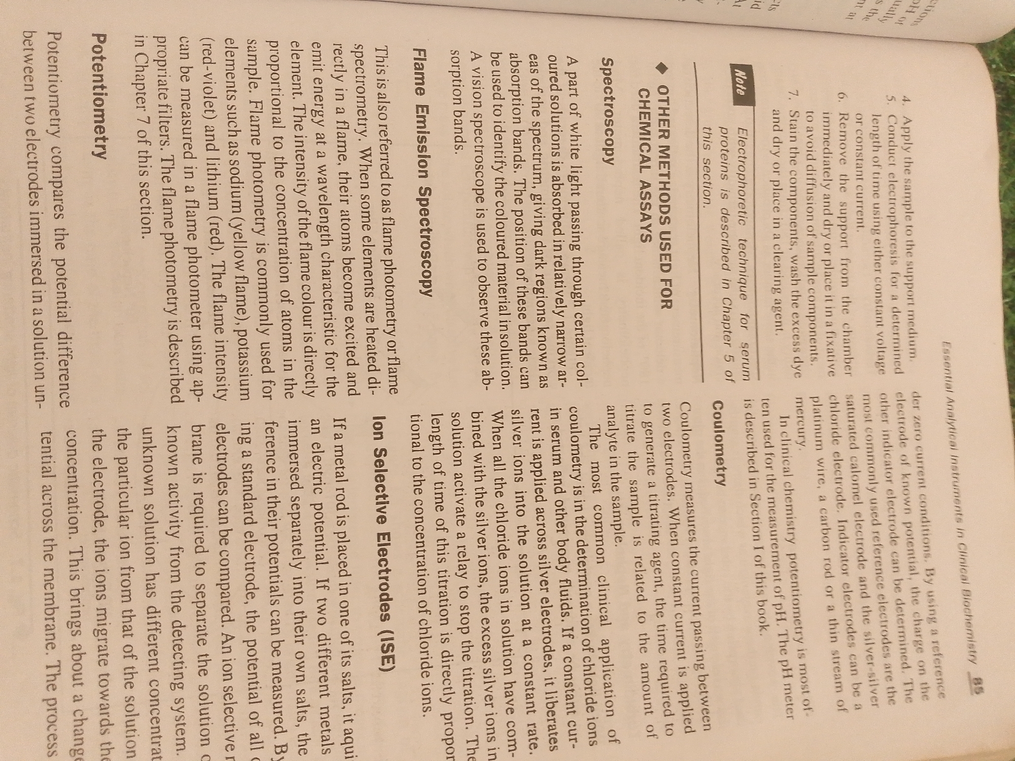 This media file is a close-up, eye-level shot of a page from a book. The text on the page is predominantly black, with some headings in bold. The subjects are the printed words and the paper they are on. The scene is a flat surface, likely a table or desk, with the book lying open.
The primary activity is reading or studying the text. The text discusses scientific concepts related to analytical instruments, chemical assays, spectroscopy, flame emission spectroscopy, potentiometry, coulometry, and ion-selective electrodes. It also includes a "Note" about electrophoretic techniques for serum proteins. There is a page number "85" visible, along with a header indicating "Essential Analytical Instruments in Clinical Biochemistry."
There are no people, objects other than the book, or discernible environmental details like time of day or weather. The location context provided (Jalingo, Nigeria) cannot be inferred from the visual information in the image itself. The text is clear and legible.