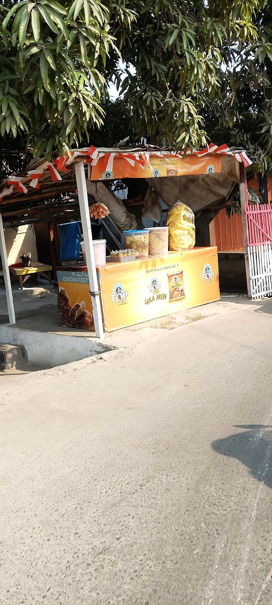 This is a full, eye-level, outdoor, slightly angled shot of a small roadside food stall. The stall is covered by a thatched roof adorned with red and white Indonesian flags. The front of the stall features a prominent yellow banner with the "TOP Coffee Gula Aren" logo and images of coffee beans and palm sugar. Displayed on the counter are several containers of food and drinks, including a large bag of yellow chips or snacks. A blue tarp is visible behind the counter. The stall is situated next to a paved road, with a tree providing shade from above. In the background, a pink and white fence can be seen. The scene appears to be in Babelan, Indonesia.