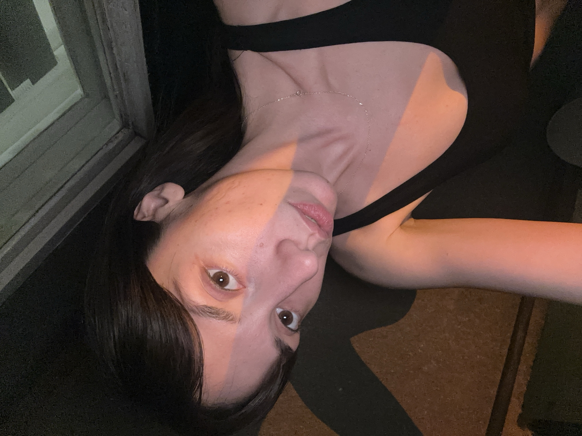 A woman is seen in a close-up, tilted selfie, with her head oriented towards the lower left of the frame. She has brown eyes and dark brown hair, wearing a black top and a delicate necklace. The background includes a partially visible window frame to the left, suggesting an indoor setting. The scene is dimly lit with a warm, orange hue on one side of her face, contrasting with the darker areas. The location is not discernible as being specifically within the Vracar Urban Municipality, Serbia, based solely on the provided image.