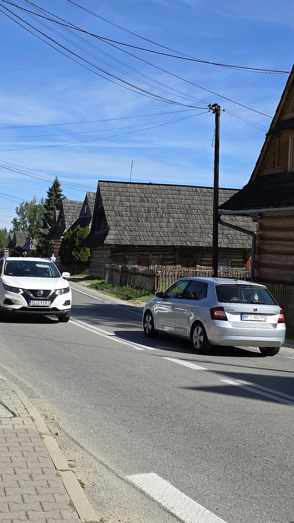 The image depicts a street scene in Chochołów, Poland, characterized by traditional wooden architecture. In the foreground, a paved road runs through the scene, with a sidewalk made of interlocking pavers on the left. Two cars are visible on the road: a white Nissan Qashqai in the left lane, and a silver Skoda Fabia in the right lane. The Nissan has a license plate reading "SLU 1YR3". The Skoda has a license plate reading "WPT 60790". The buildings lining the street are single-story wooden structures with steep, shingled roofs, typical of the region's folk architecture. A wooden fence runs along the front of the properties. Overhead, power lines crisscross the clear blue sky, supported by a wooden utility pole. The sun is shining, casting shadows on the road. A single individual, a man wearing a light blue shirt, is visible walking on the sidewalk in the distance, between the two cars. The overall impression is of a quiet, rural street in a historic village.