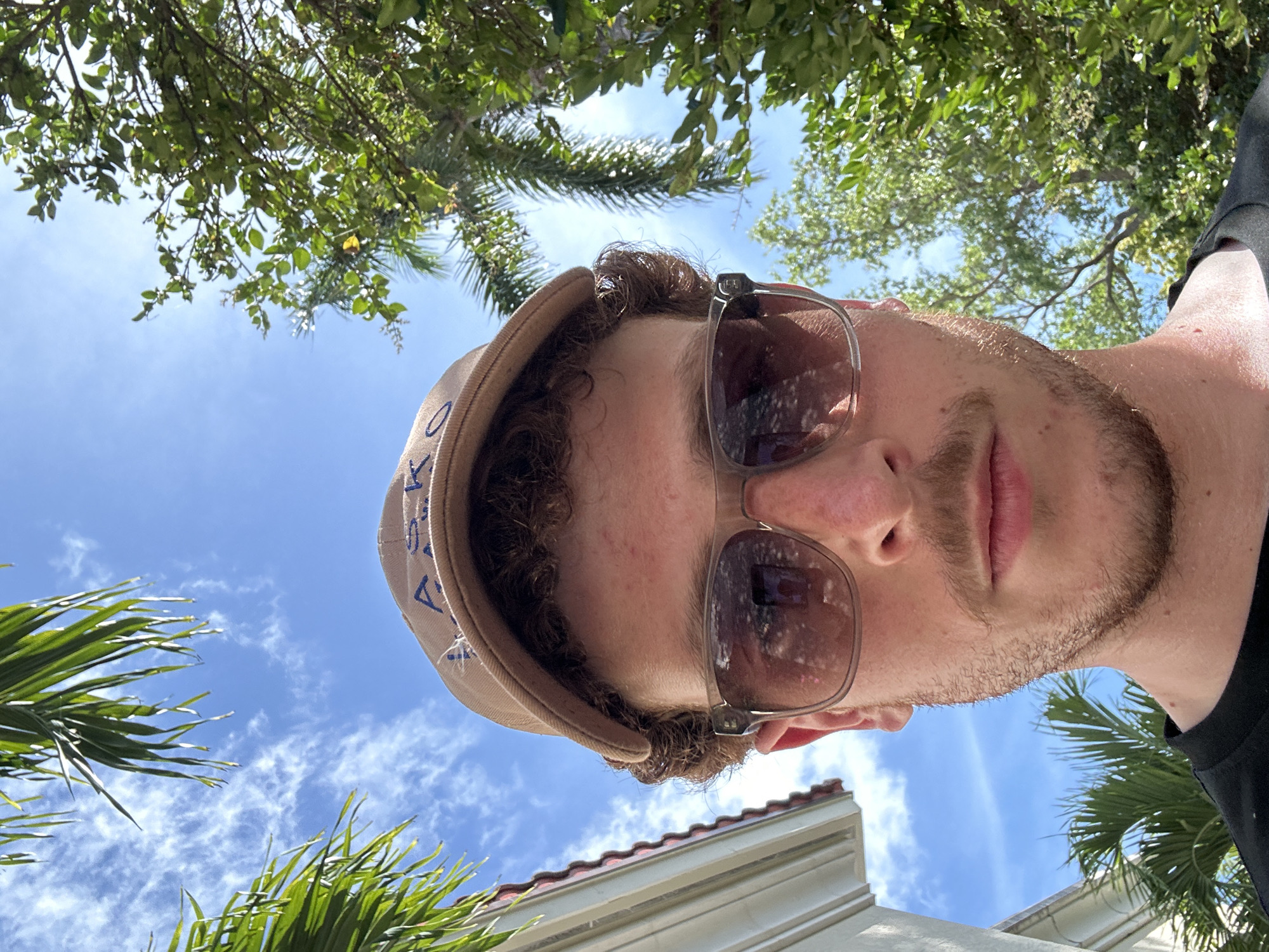 A low-angle, outdoor selfie captures a man in Aventura, Florida, United States. The man is wearing a brown cap and sunglasses. He has fair skin and a light beard. The background consists of a blue sky with scattered clouds, trees, and a glimpse of a building. Palm trees are visible on the left side and bottom right corner of the image. The building has a white wall and a red tile roof. Sunlight is present, creating shadows and reflections in the sunglasses.