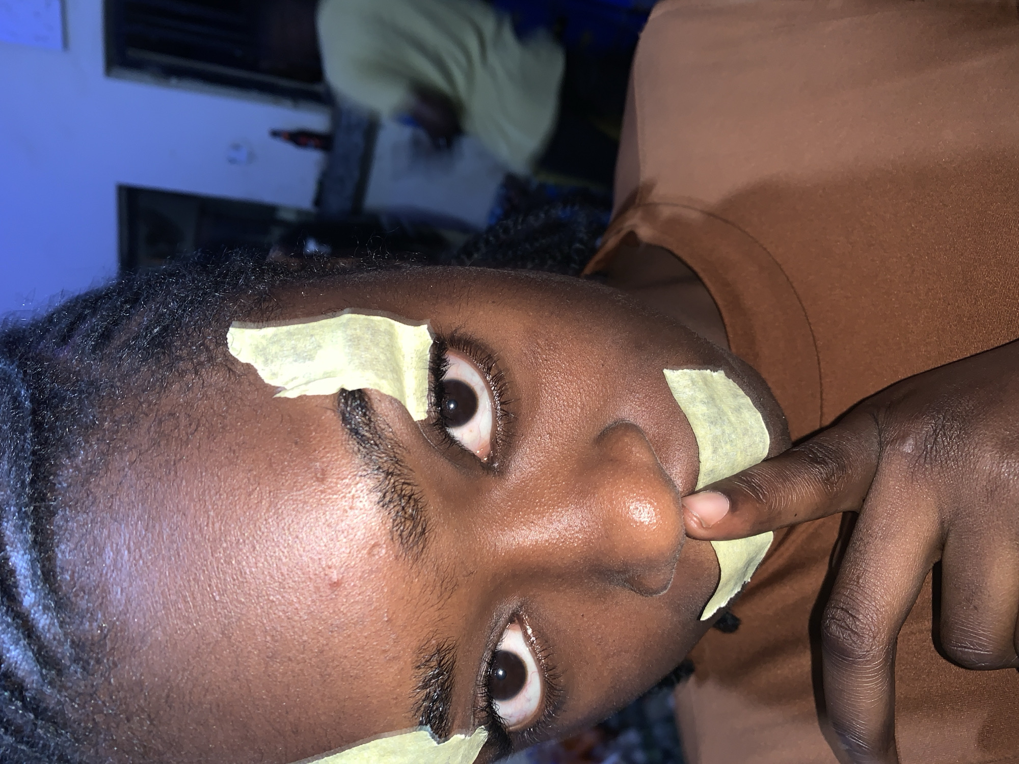 This image, captured in the city of New Stadium, Nigeria, features a close-up portrait of an individual, likely female, with dark skin and dark braided hair, directly facing the camera. The person is wearing a brown, seemingly satin-like, high-neck top. Several light yellow/green paper-like strips are adhered to their face: one above the right eyebrow, another to the left of the left eye, and a third horizontally across the mouth. The individual's right index finger is pressed vertically against the paper strip covering the mouth, creating a "shhh" gesture. Both eyes are open and directly engaging the viewer; the sclerae exhibit minor reddish vascularity. A small, raised epidermal blemish is visible on the forehead, slightly above the right eyebrow.
The background is out of focus. To the right, a wall is visible, illuminated by a distinct blue light source, giving it a cool, azure tint. Below this blue-lit section, a dark counter or surface is present, upon which a dark bottle, possibly glass, with a red label is situated. Further to the right and significantly blurred, the back of another individual, wearing a light yellow or green t-shirt and dark trousers, is partially visible. Two dark, rectangular doorframes or openings can be discerned in the blurred background. The overall scene suggests an indoor, casual setting with mixed lighting conditions.