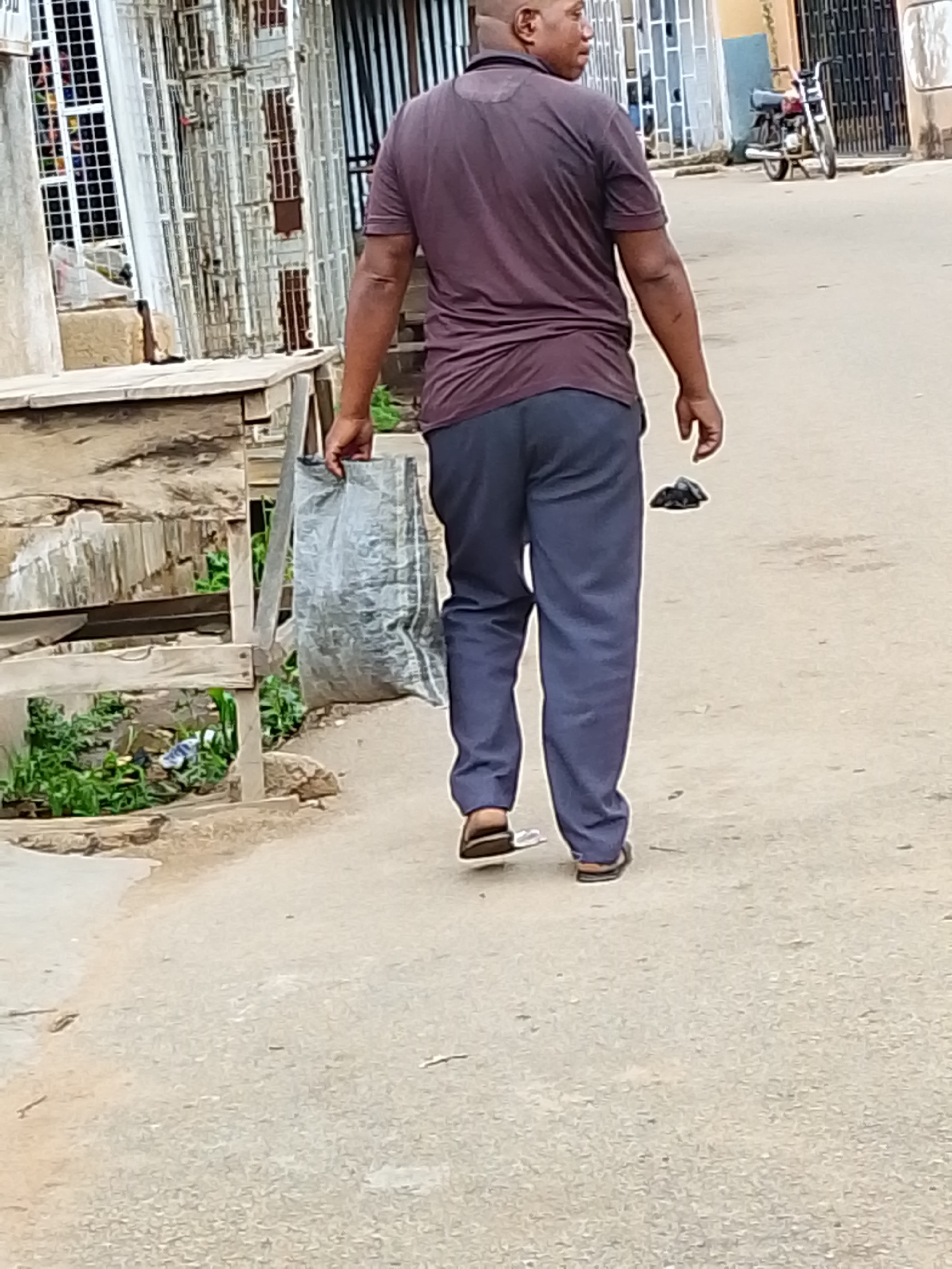 A man is walking away from the camera on an urban street. He is carrying a silver plastic bag in his left hand. The man is wearing a maroon short-sleeved shirt, blue trousers, and sandals. In the background, there is a building with a white wrought-iron security gate. A motorcycle is parked on the side of the road further down the street. There is a small wooden structure to the left of the man. A dark object lies on the ground in front of him. The street appears to be paved and is light gray in color. Greenery is visible near the wooden structure.