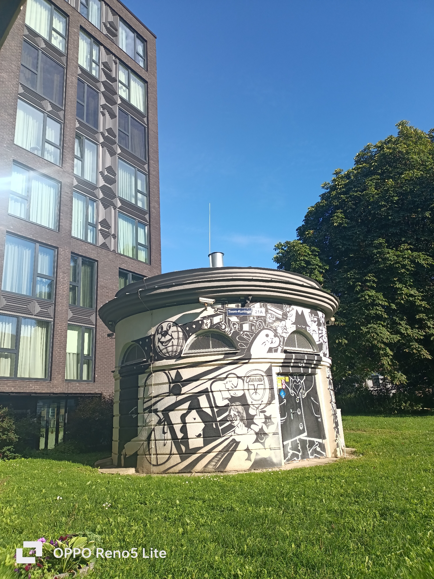 This image depicts a cylindrical building covered in black and white graffiti, located in Tallinn, Estonia. The building is situated on a well-maintained lawn, with a tall, modern brick building visible to its left and a large, leafy tree to its right. The sky is clear blue, indicating a sunny day. The graffiti on the circular building features various cartoonish figures and abstract designs. A street sign reading "Toom-Kuninga 21A" is affixed to the upper portion of the building's facade. The scene is devoid of people.